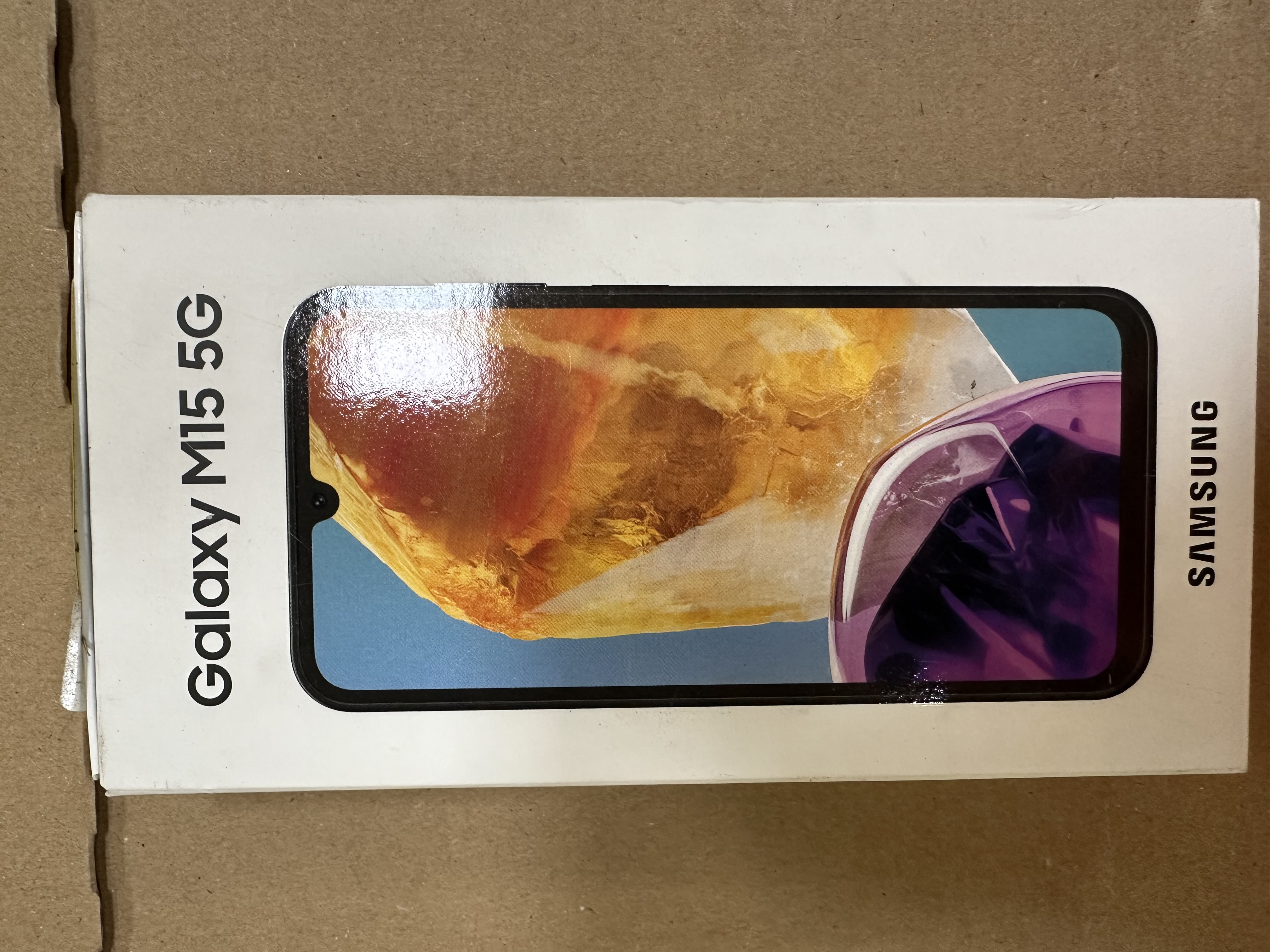 Pudełko telefonu Galaxy M15 5G firmy Samsung. Ekran telefonu jest widoczny z kolorowym abstrakcyjnym obrazem.