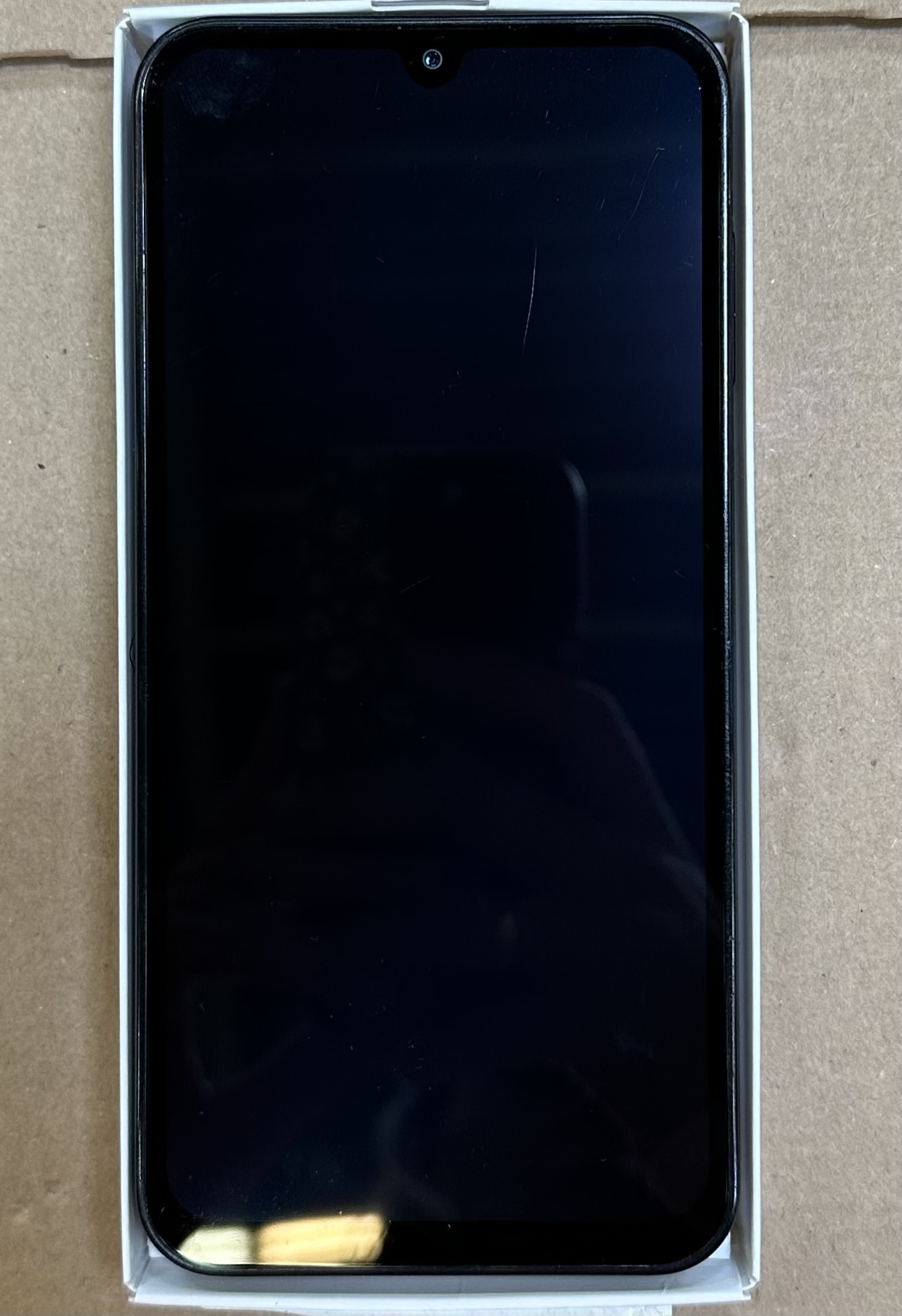 Czarny smartfon jest w białym pudełku. Ekran jest czarny z przednim aparatem u góry. Tło jest brązowe.