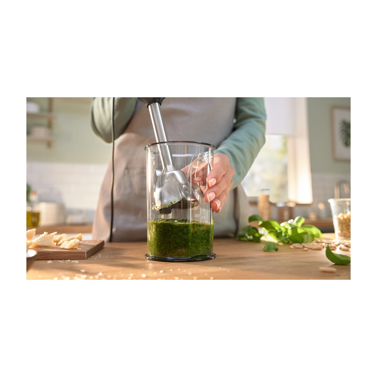 Un frullatore a immersione in un barattolo di vetro con pesto verde. Un tavolo di legno e una cucina.