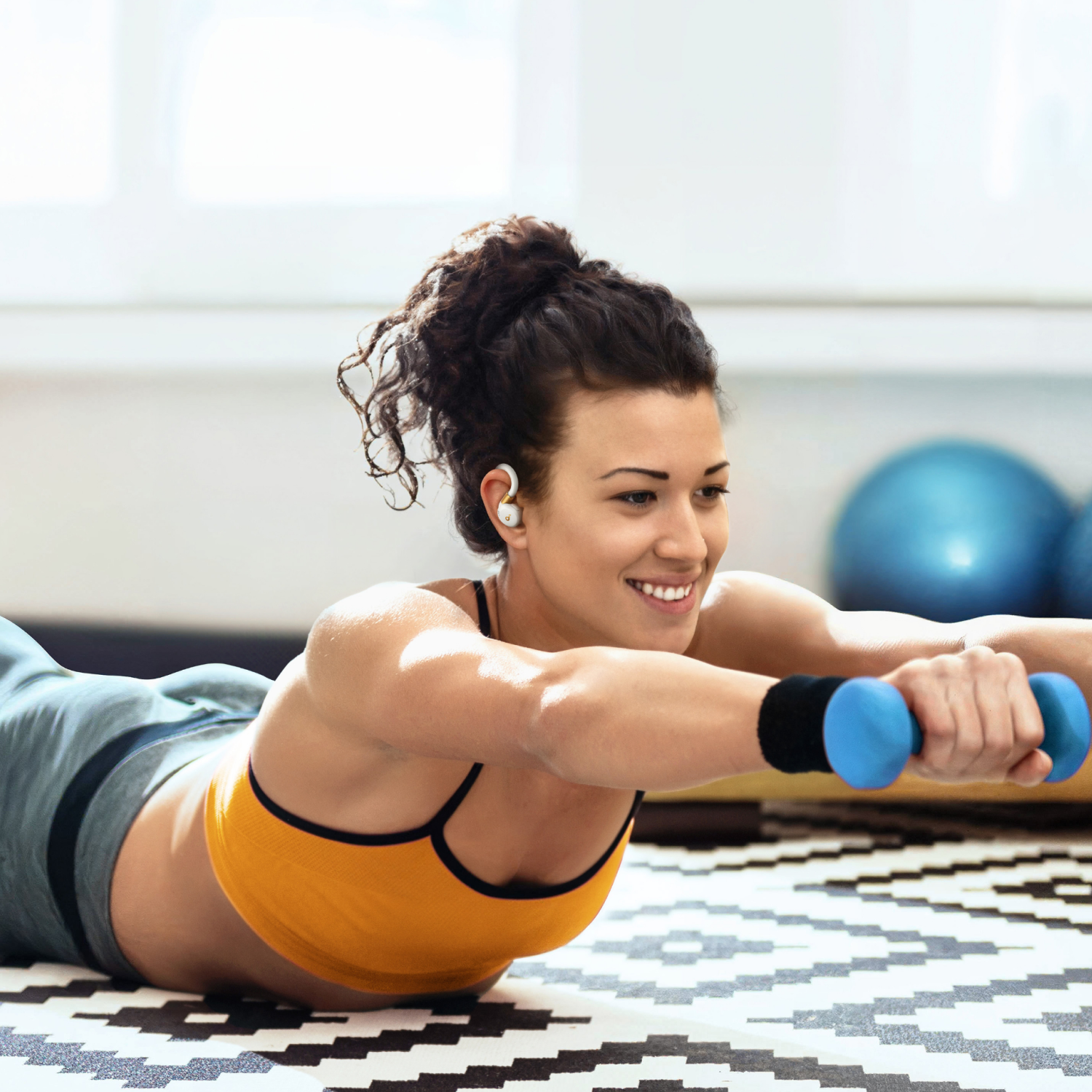 Woman exercising with dumbbells, wearing earbuds with gold accents. She is smiling and on a mat.
