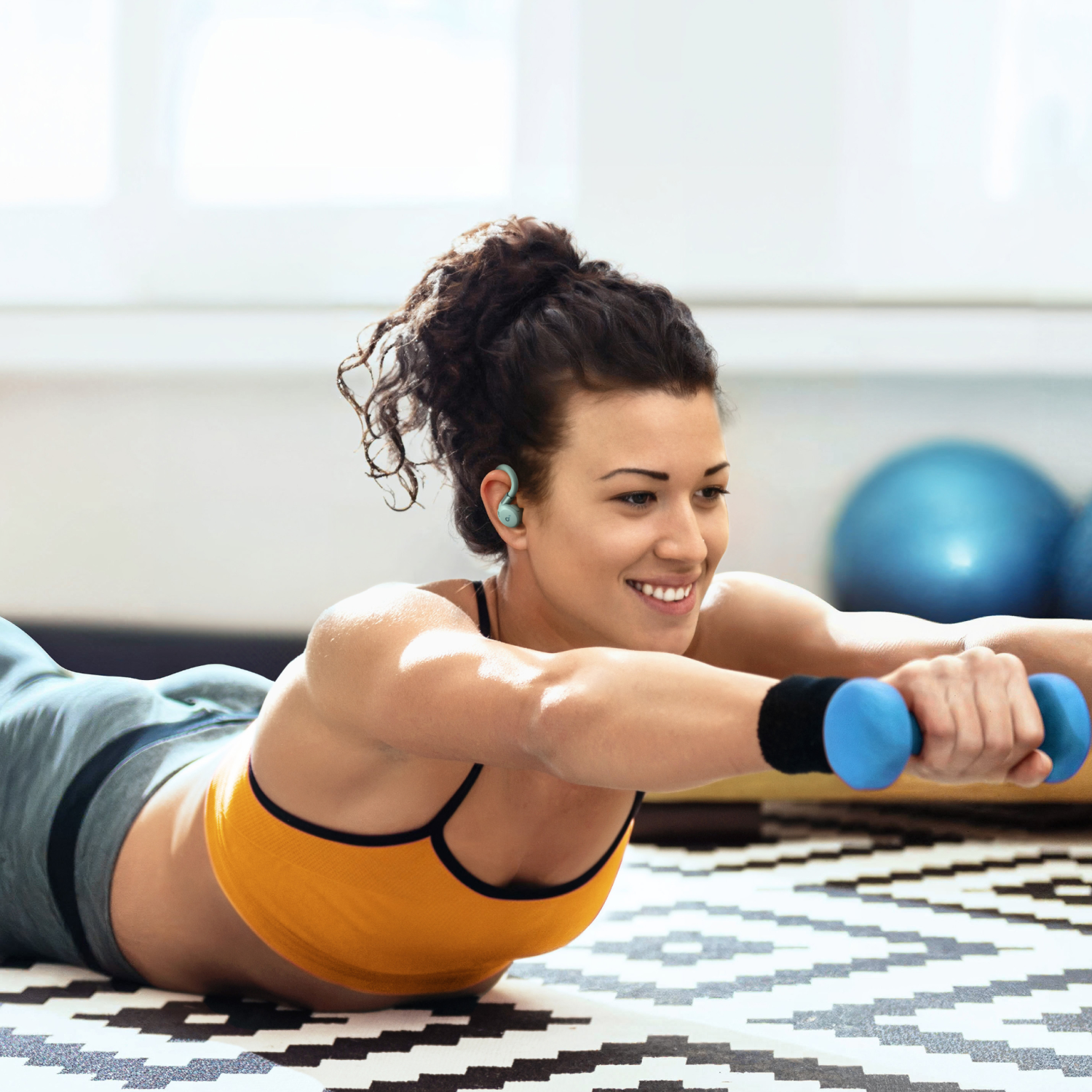 A woman in activewear exercises. She wears green earbuds, smiles, and holds dumbbells.