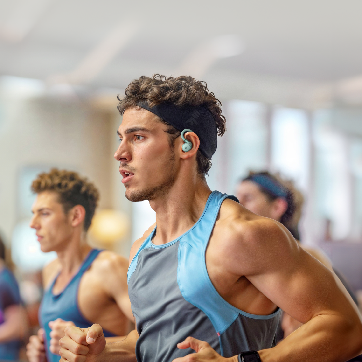 A man runs in a gym wearing earbuds. Other people are in the background.