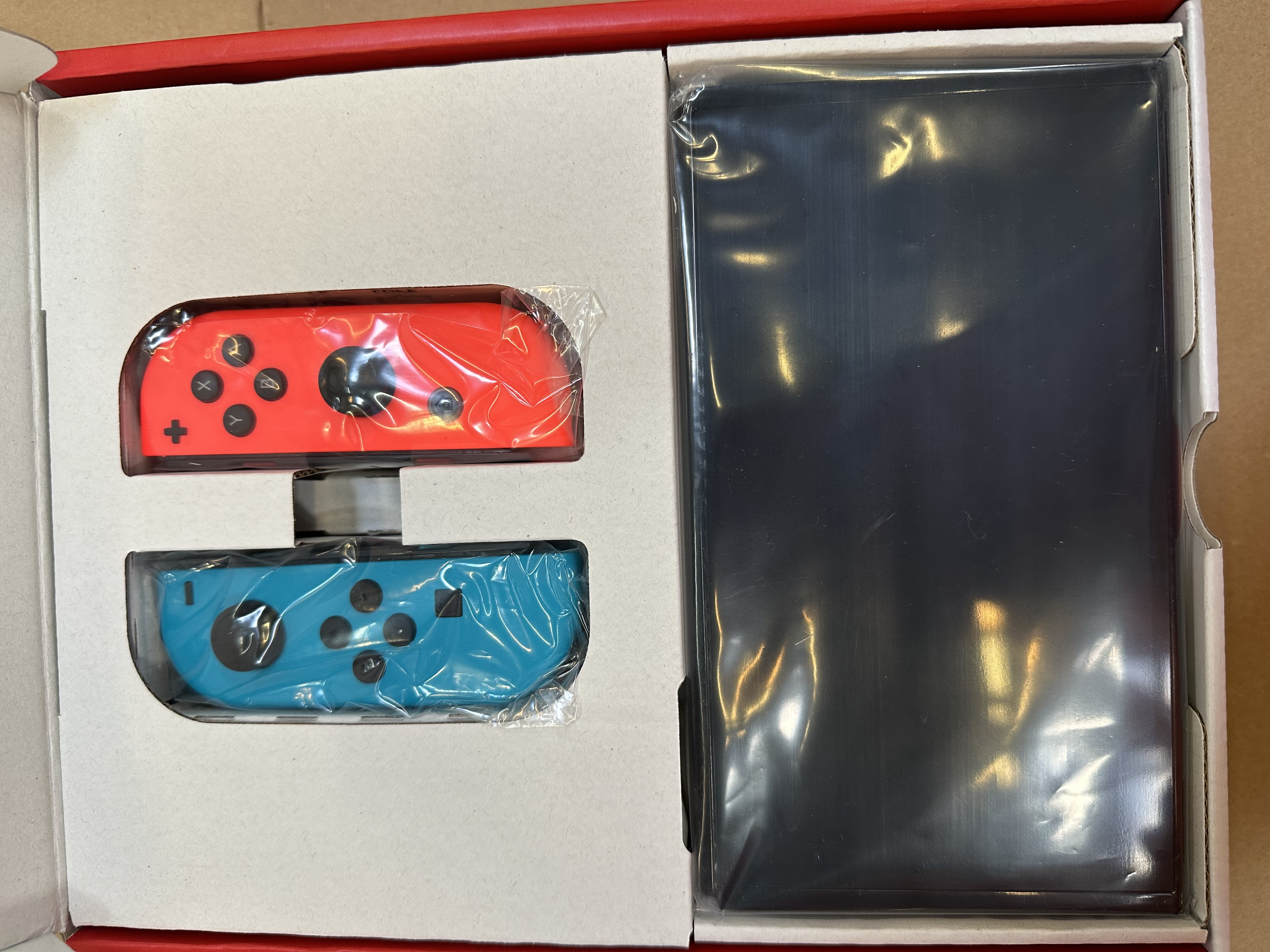 Otwarty karton Nintendo Switch. Czerwone i niebieskie kontrolery, czarna konsola, wszystko zapakowane.