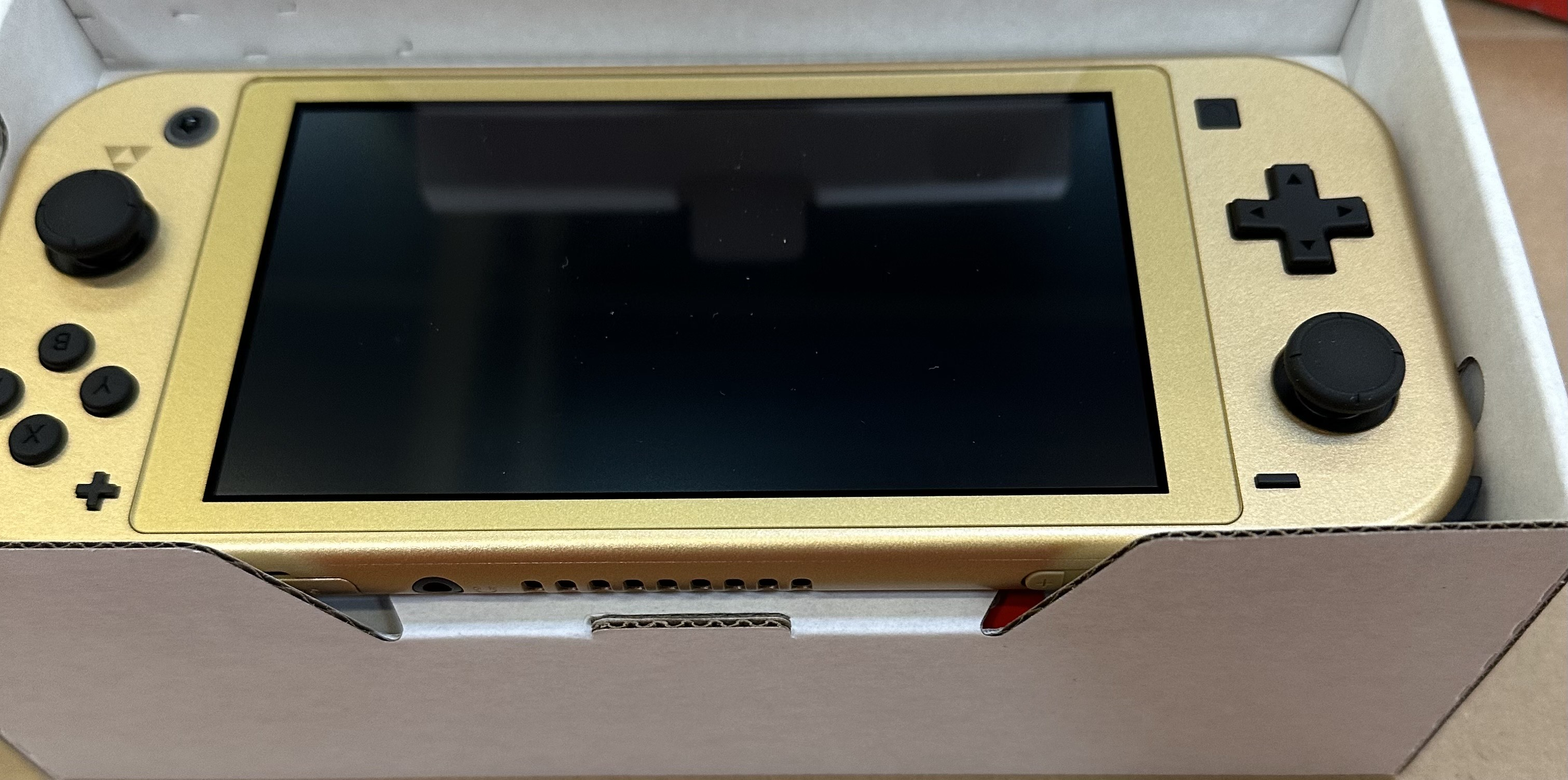 Złoty Nintendo Switch Lite w opakowaniu, widoczny z góry. Ekran jest czarny.