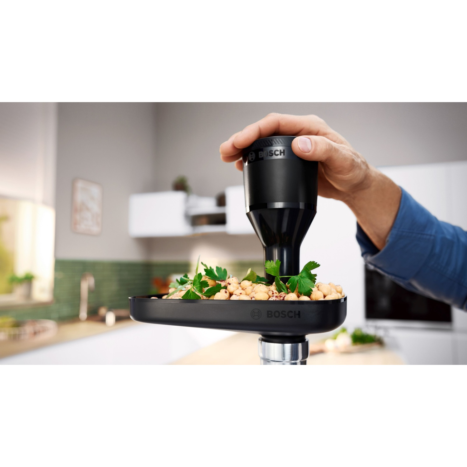Una mano usa un robot da cucina Bosch nero per elaborare il cibo. Ceci e prezzemolo sono visibili nel vassoio nero.