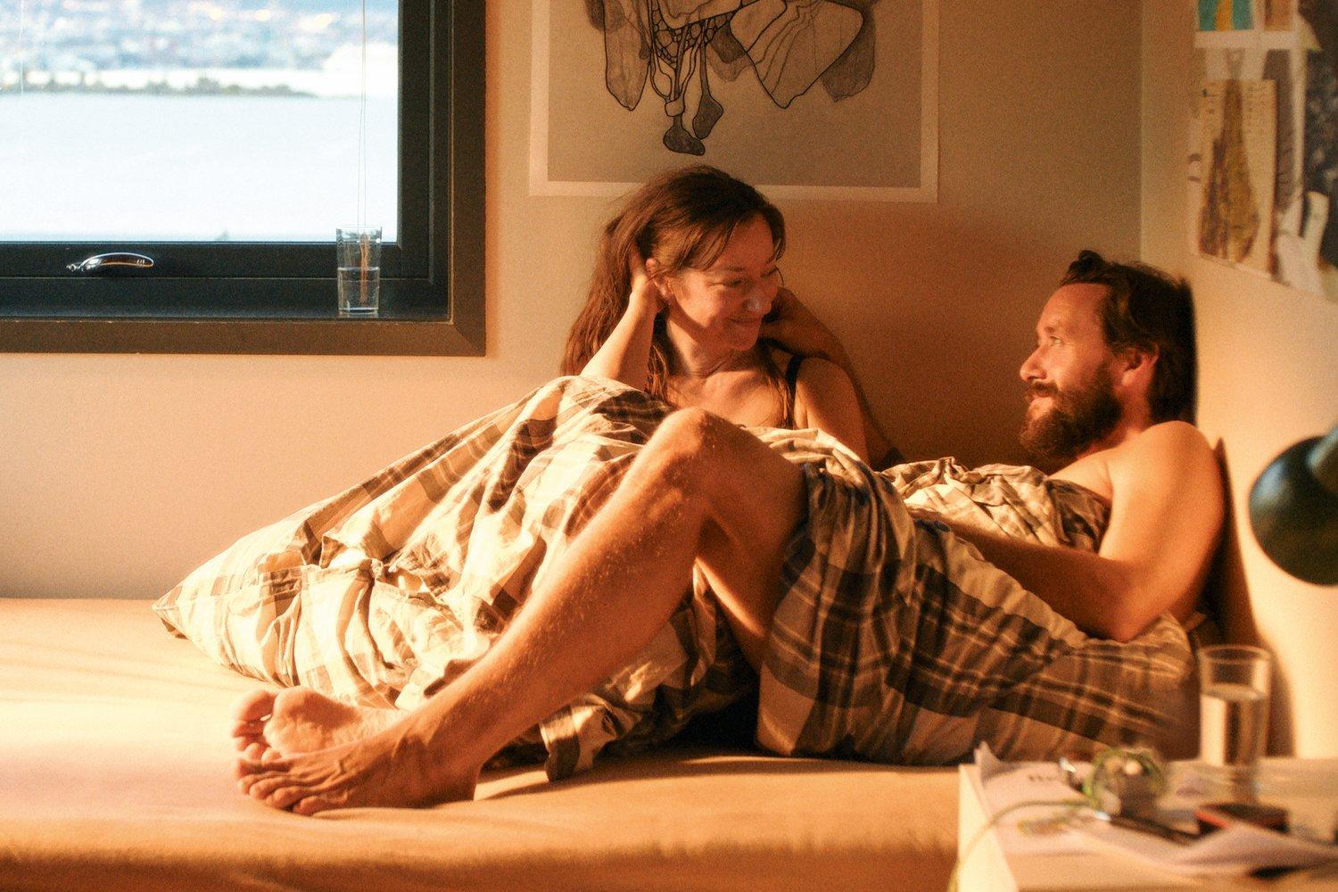 Ein Paar liegt im Bett. Sie sind unter einer Decke. Ein Glas Wasser ist sichtbar.