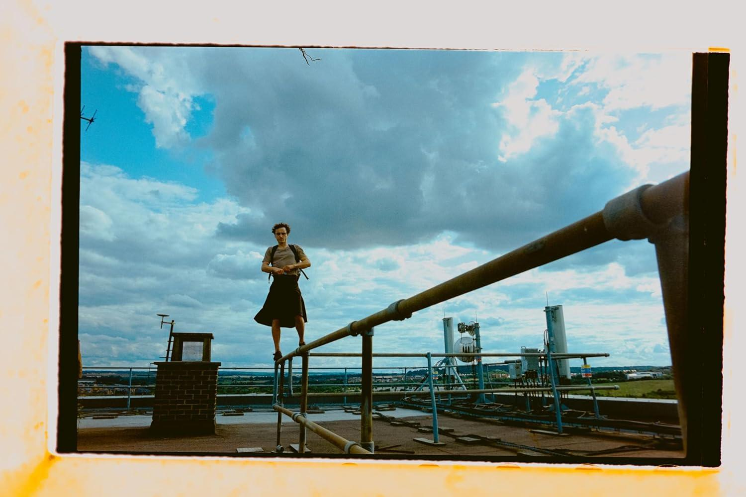 Ein Mann steht auf einem Dachgeländer mit blauem Himmel und Wolken. Antennen und Gebäude sind sichtbar.