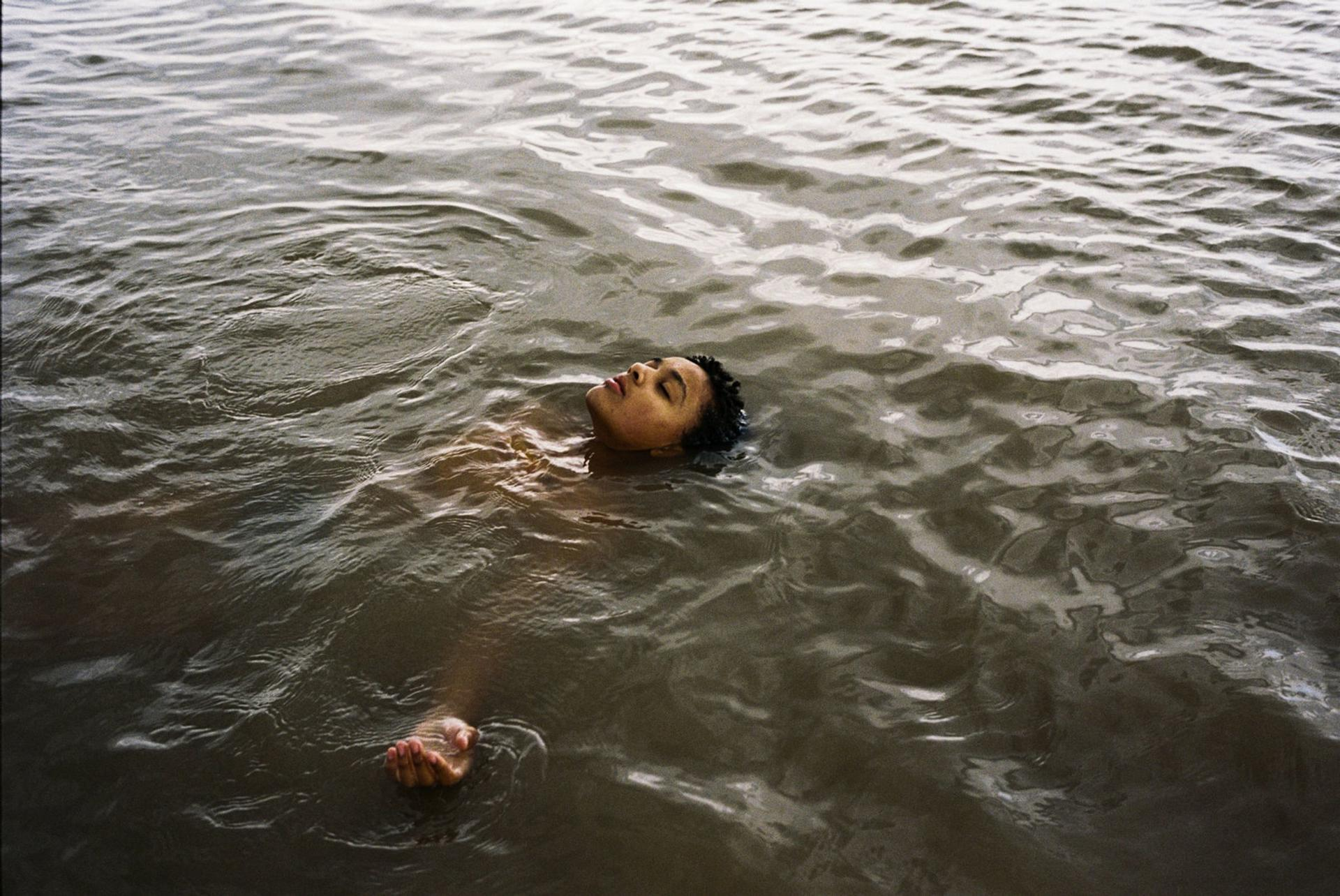 Person schwimmt im Wasser, Gesicht nach oben. Ruhiges Wasser, bedeckter Himmel. Arm und Hand sichtbar.