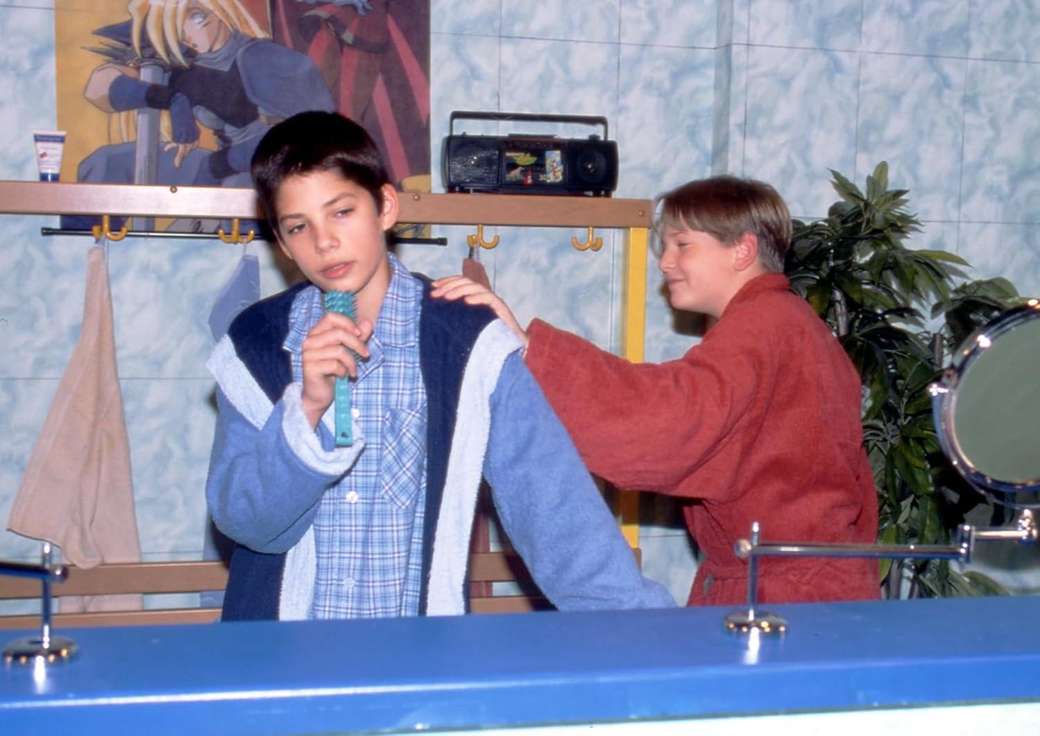 Zwei Jungen in einem Badezimmer. Einer hält ein Mikrofon und der andere legt etwas auf seine Schulter.