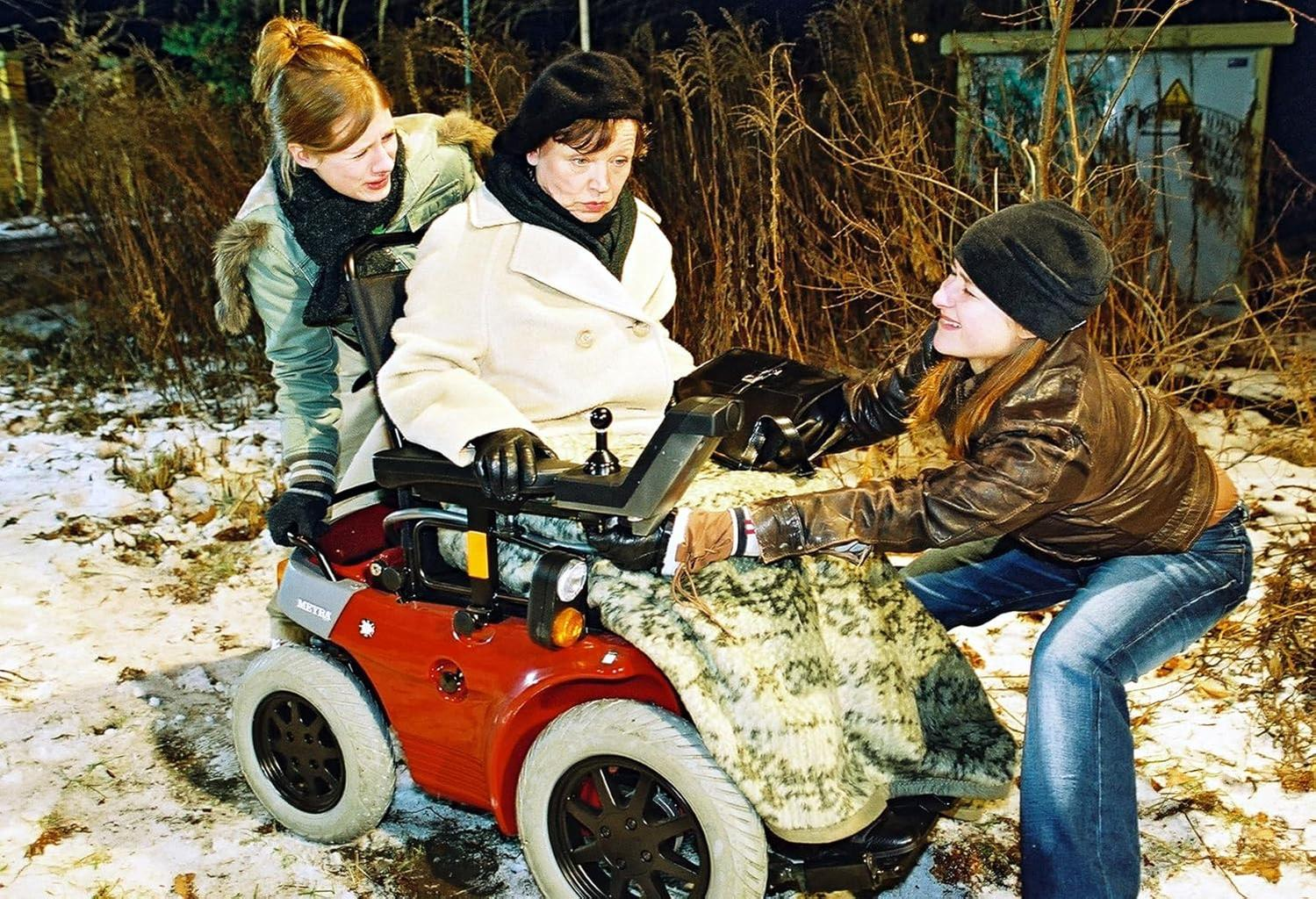Drei Frauen um einen roten Motorrollstuhl. Eine sitzt. Verschneite Umgebung im Freien.
