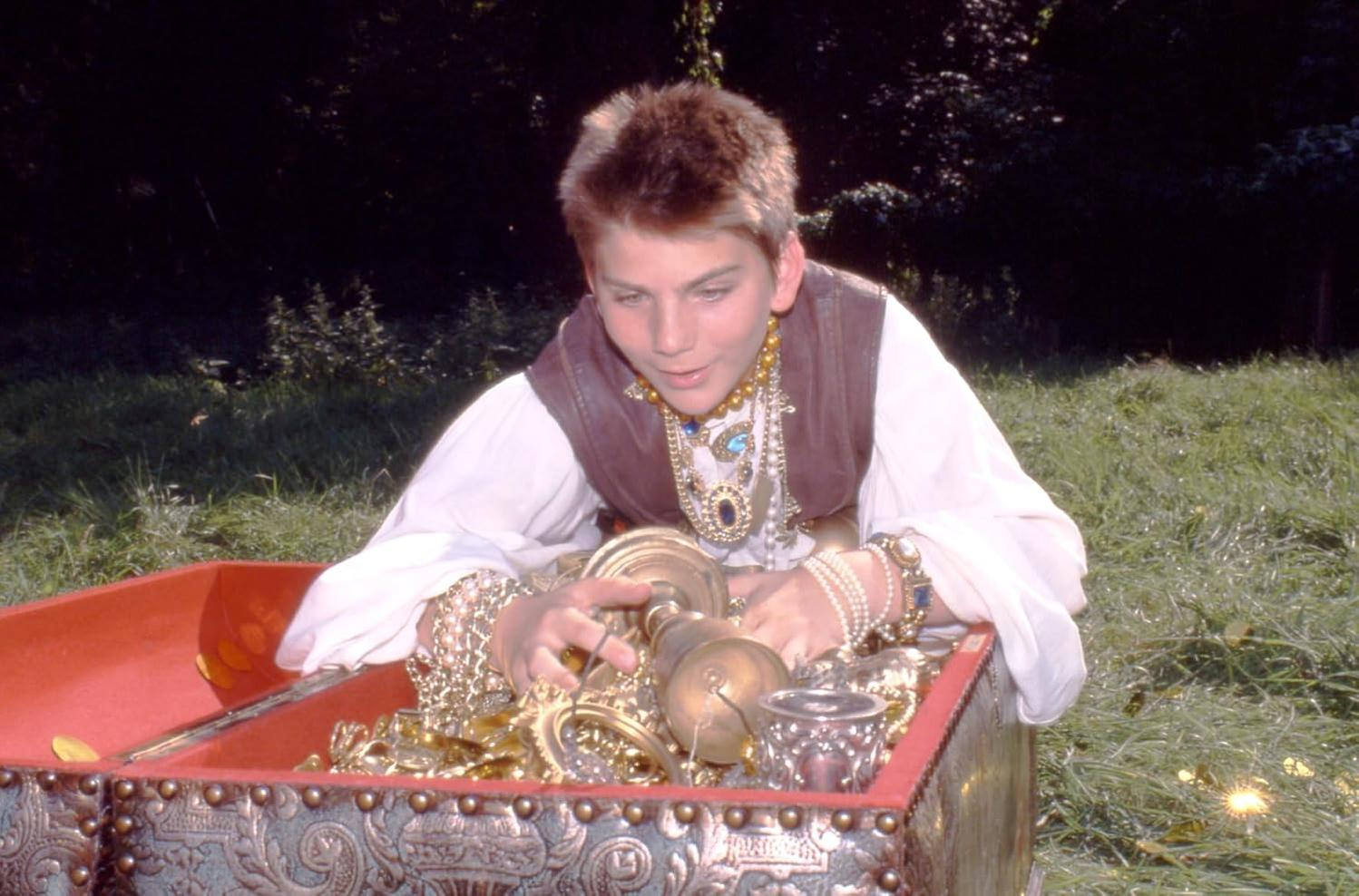 Junger Mann in einer Schatzkiste mit Goldschmuck. Truhe ist offen. Grüner Gras Hintergrund.