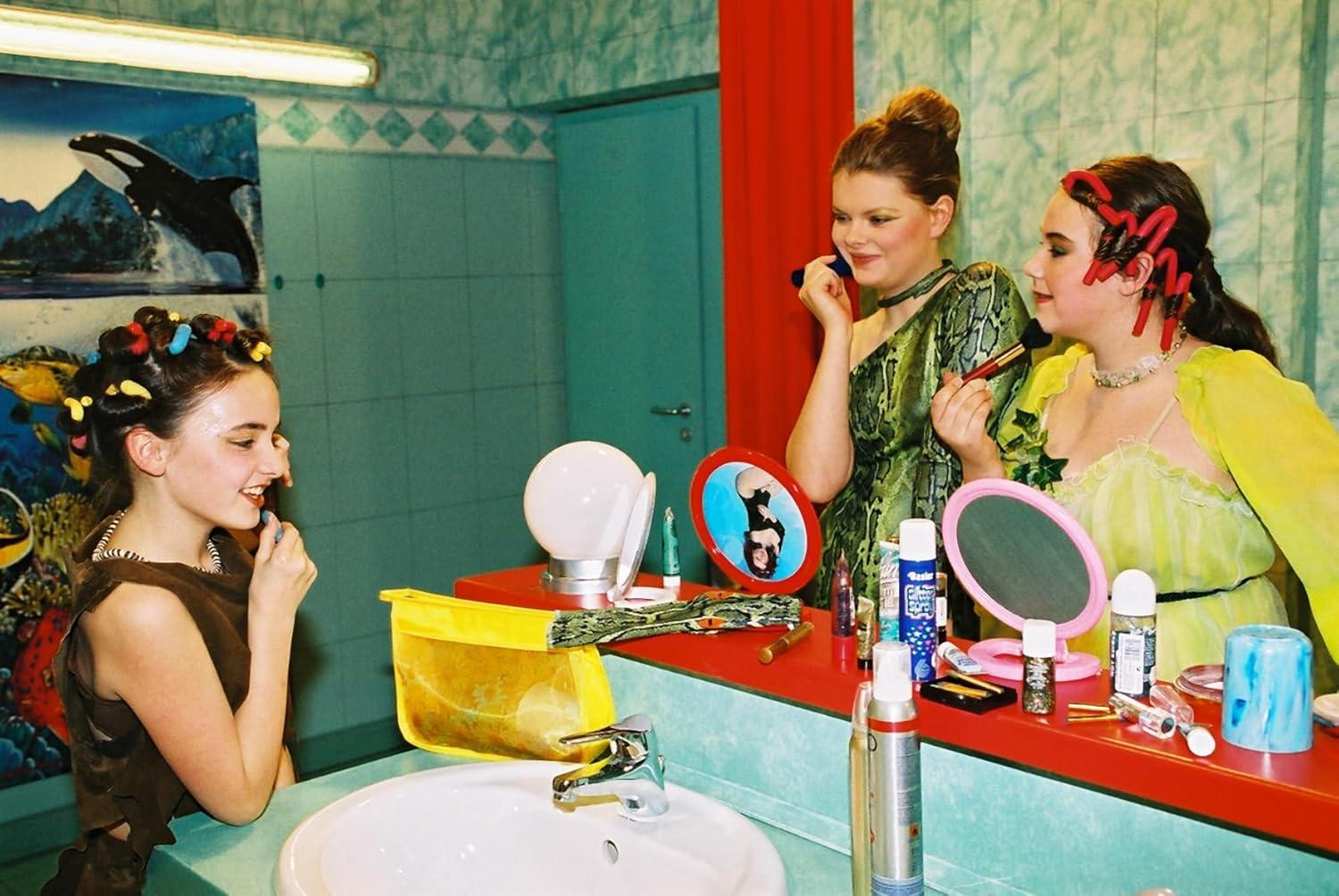 Drei Mädchen in einem Badezimmer. Eines schminkt sich. Spiegel, Lichter und Kosmetika sind vorhanden.