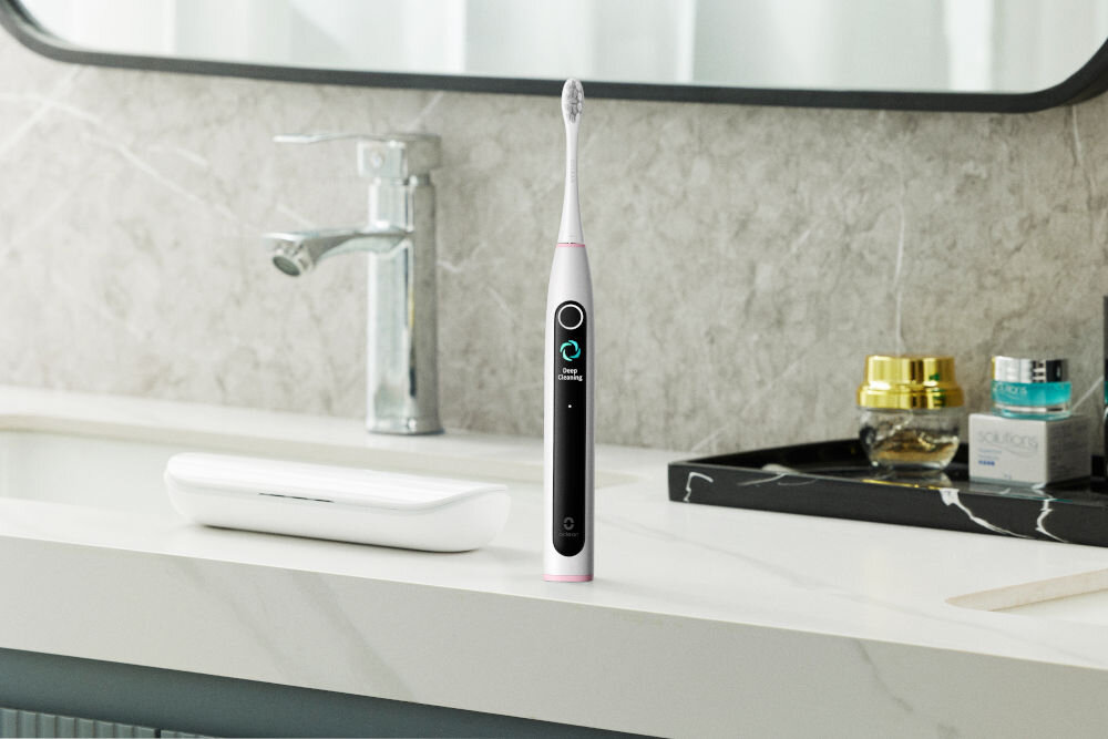 Elektryczna szczoteczka do zębów stoi na blacie w łazience. Obecne są również etui, kran i kosmetyki. Tło to marmurowa ściana.