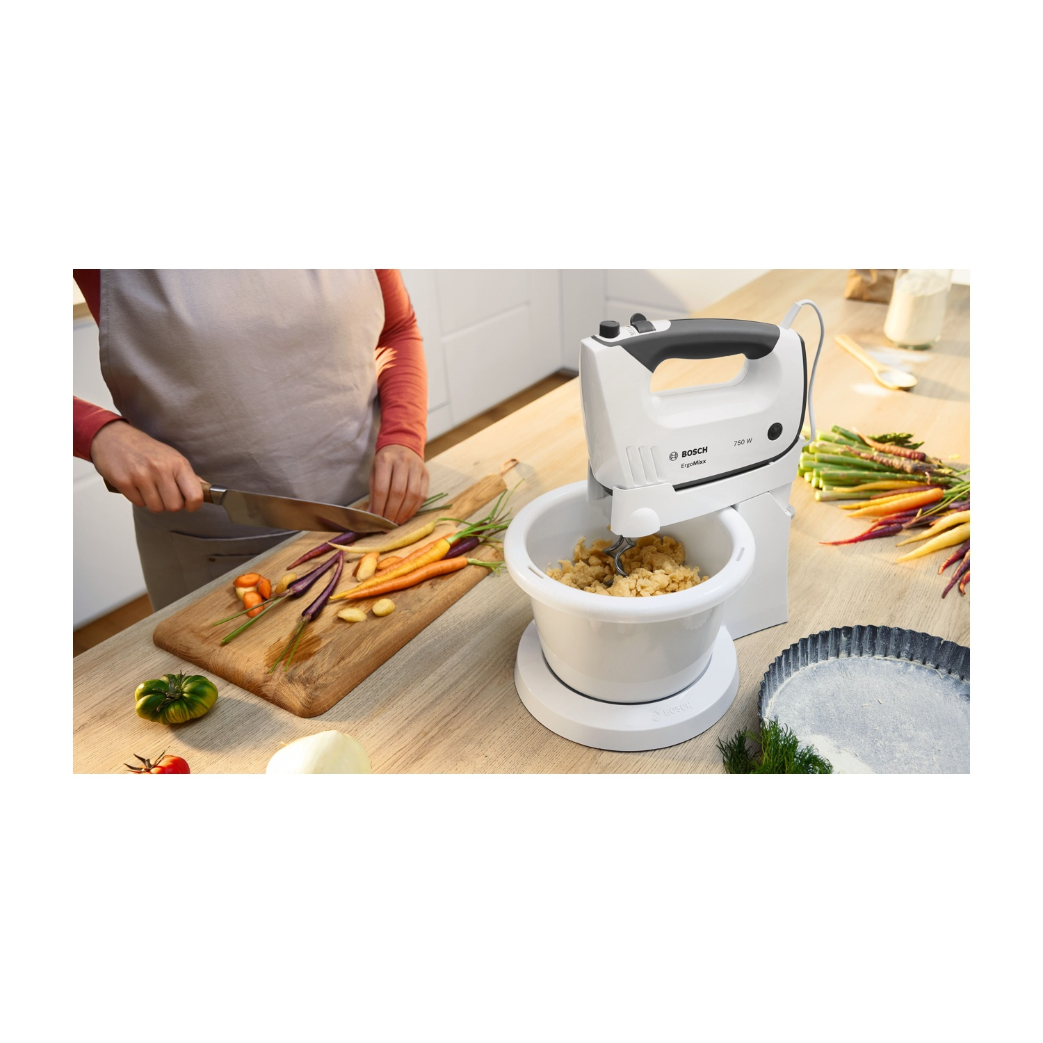 Donna che usa un frullatore a mano con carote e verdure su un tagliere. Ambiente cucina.