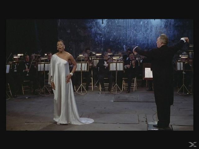 Eine Frau singt auf einer Bühne. Ein Orchester und ein Dirigent sind im Hintergrund.