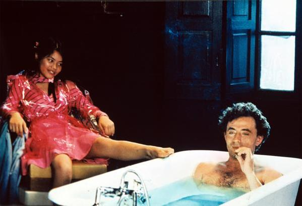 Eine Frau in rosa Kleid sitzt, während ein Mann in der Badewanne raucht.