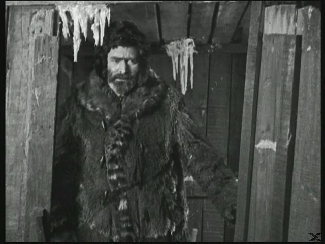 Mann in einem Pelzmantel, der in einem hölzernen Türrahmen steht. Eis hängt von oben. Schwarz und Weiß.
