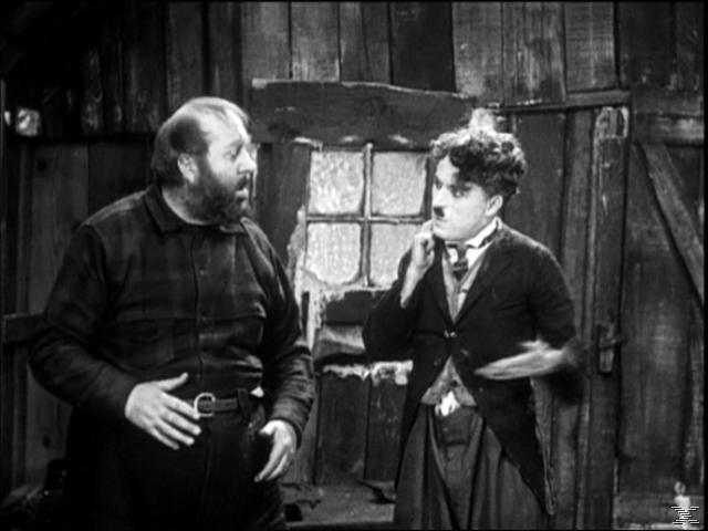 Chaplin und ein anderer Mann unterhalten sich. Chaplin hat einen Schnurrbart und trägt einen Anzug. Schwarzweiß.