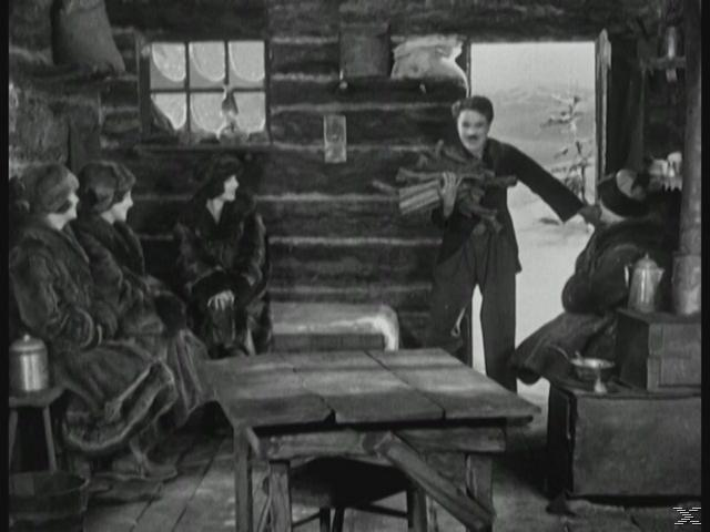 Ein Mann betritt mit Holz in der Hand eine Hütte mit sitzenden Frauen. Tisch und Ofen sind im Raum.