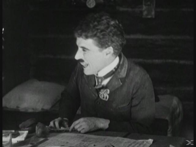 Ein lächelnder Mann mit Schnurrbart sitzt an einem Tisch. Papiere und ein Becher sind vorhanden. Dunkler Hintergrund.