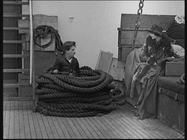 Mann in einem aufgerollten Seil, Frau sitzt daneben auf einem Schiffsdeck. Kisten und Treppen sind sichtbar.