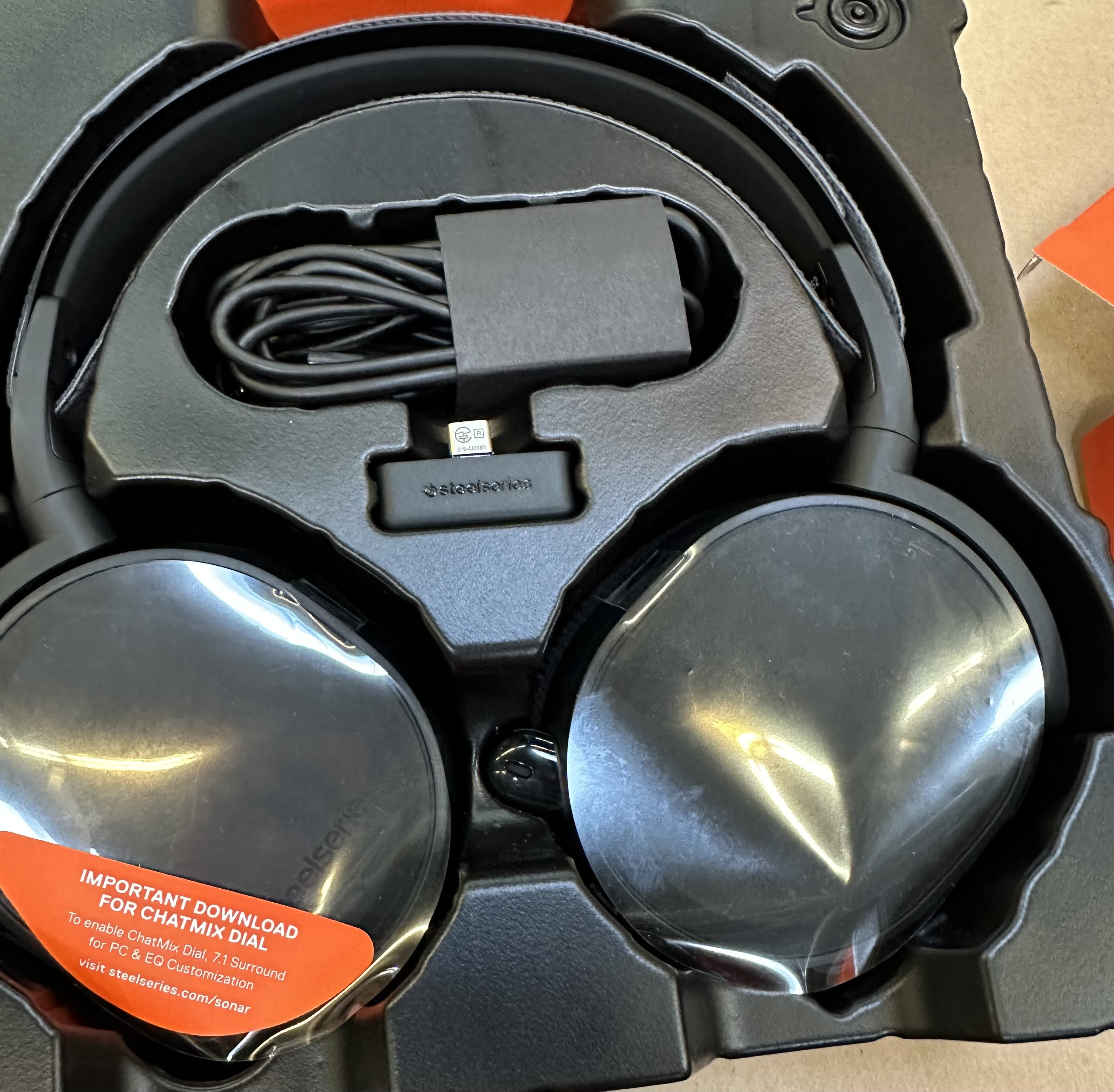 Elementy zestawu słuchawkowego SteelSeries Arctis 7+ w opakowaniu, czarne słuchawki, kabel USB i adapter.