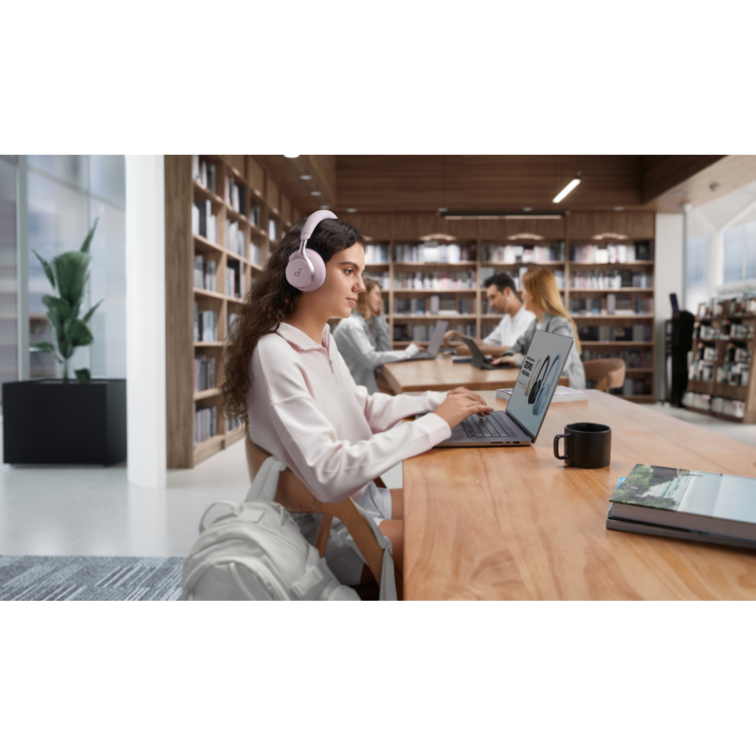 Vrouw met koptelefoon gebruikt laptop in bibliotheek. Boekenkasten en andere mensen op de achtergrond.