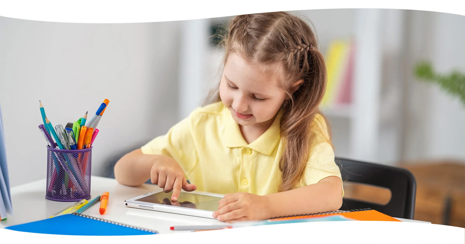 Młoda dziewczyna siedzi przy biurku, używając tabletu. Długopisy i notatniki leżą obok niej.