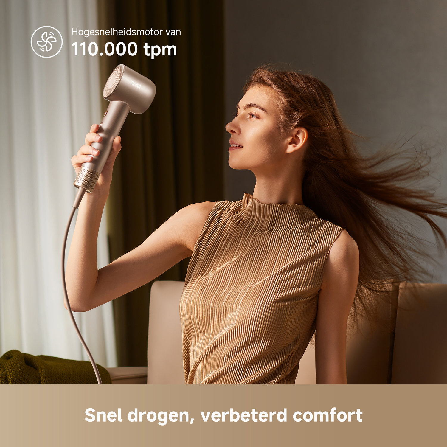 Een vrouw houdt een föhn vast, haar haar waait. De föhn is zilverkleurig. De achtergrond toont een kamer met een bank en gordijnen.