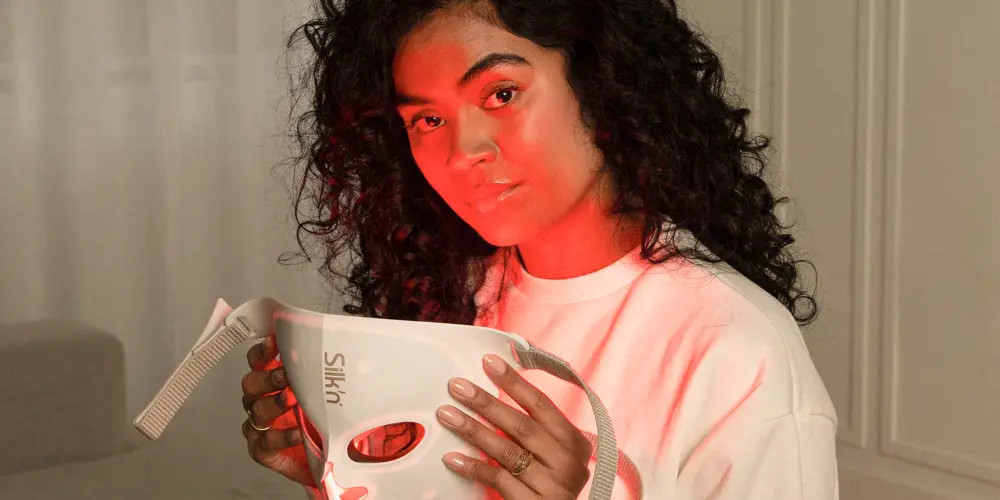 Kobieta trzyma białą maskę do twarzy oświetloną czerwonym światłem. Ma kręcone włosy i nosi białą koszulę.