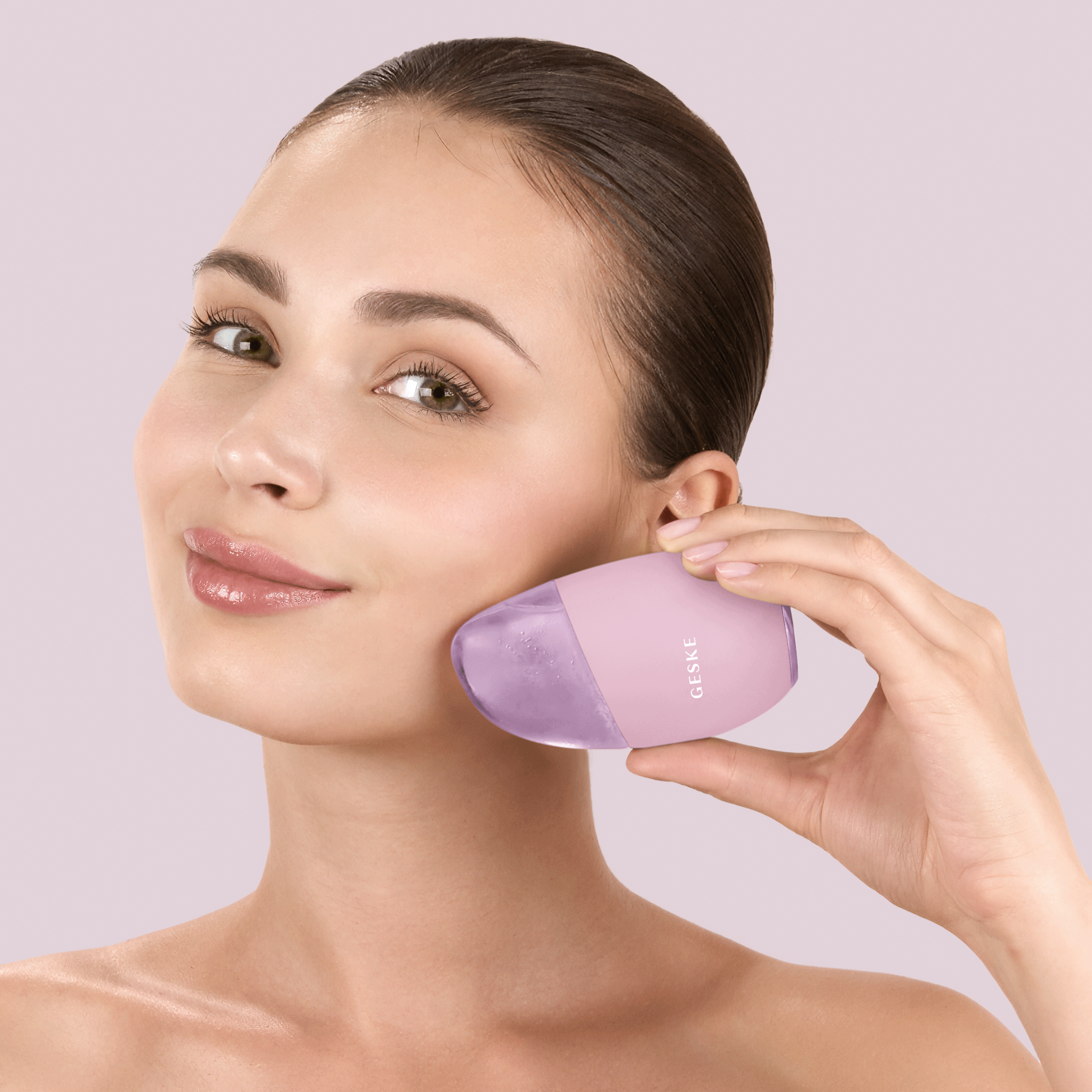 Kadın, yanağına pembe ve mor bir Geske cihazı tutuyor. Gülümsüyor. Arka plan yumuşak pembe.