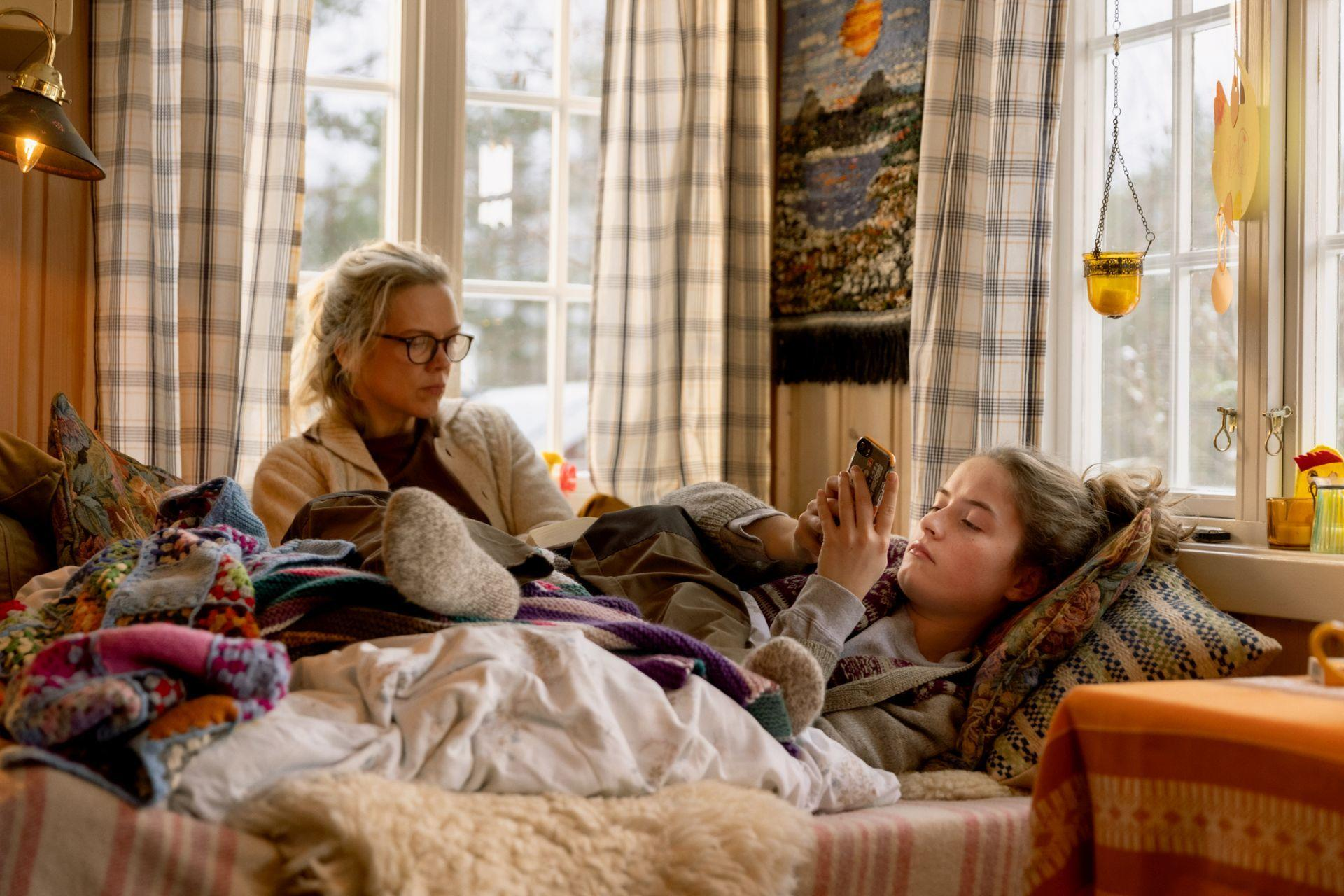 Zwei Frauen in einem Raum. Eine benutzt ein Telefon, die andere schaut sie an. Kissen und Decken auf dem Bett.