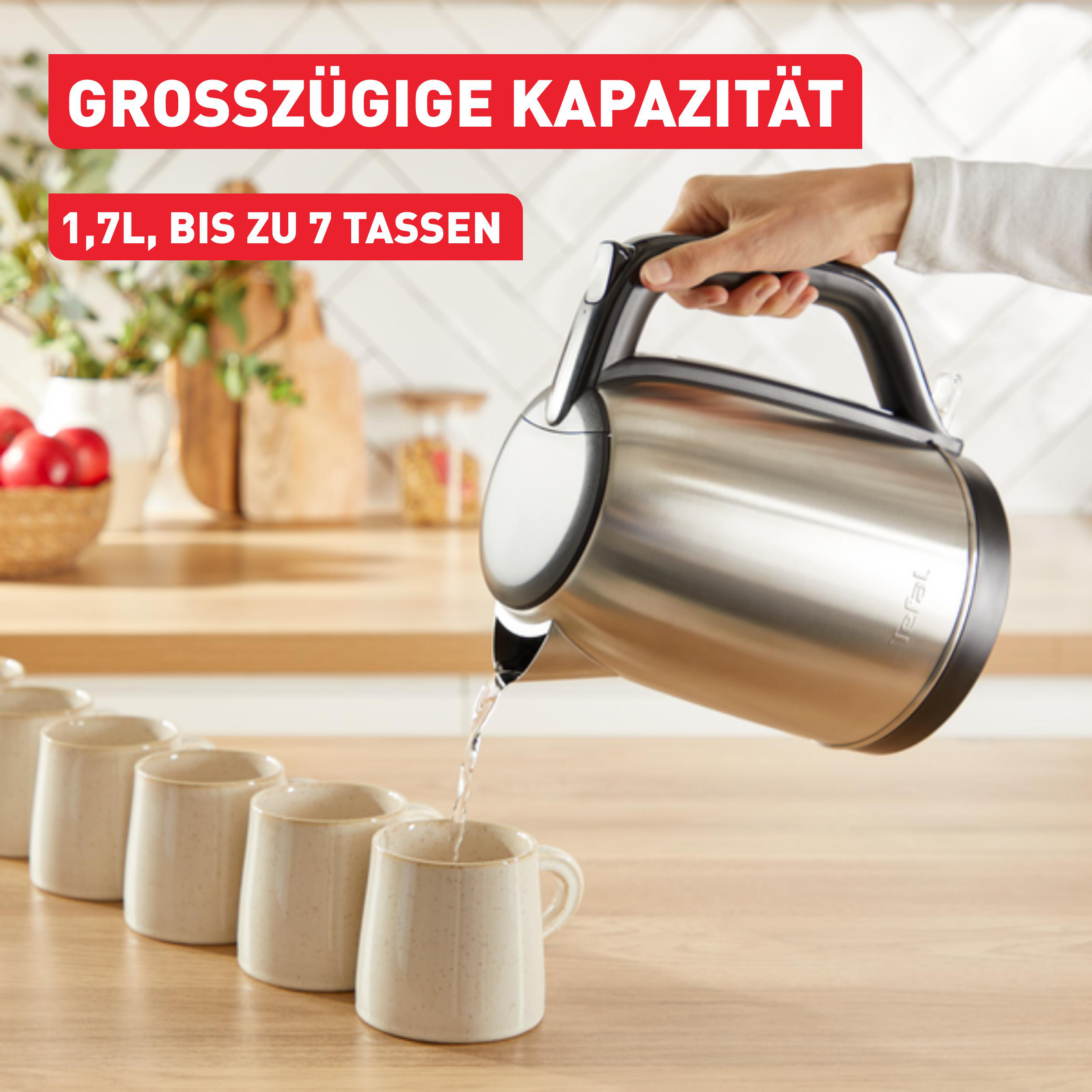Eine Hand gießt Wasser aus einem Tefal-Edelstahlkessel in Tassen auf einer Holztheke.