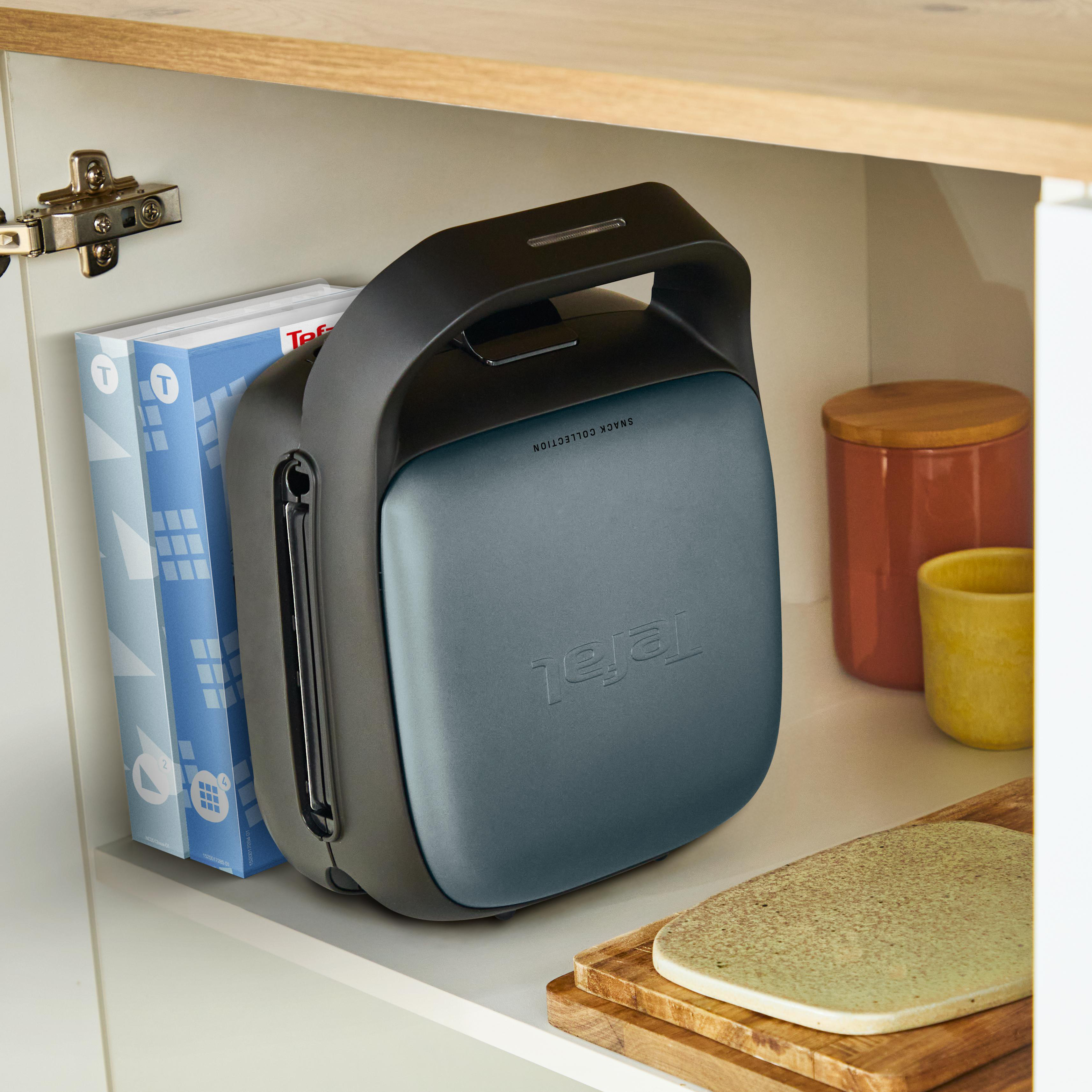 Ein blaugrüner und schwarzer Waffeleisen steht in einem Schrank mit Büchern und einem Holzschneidebrett.