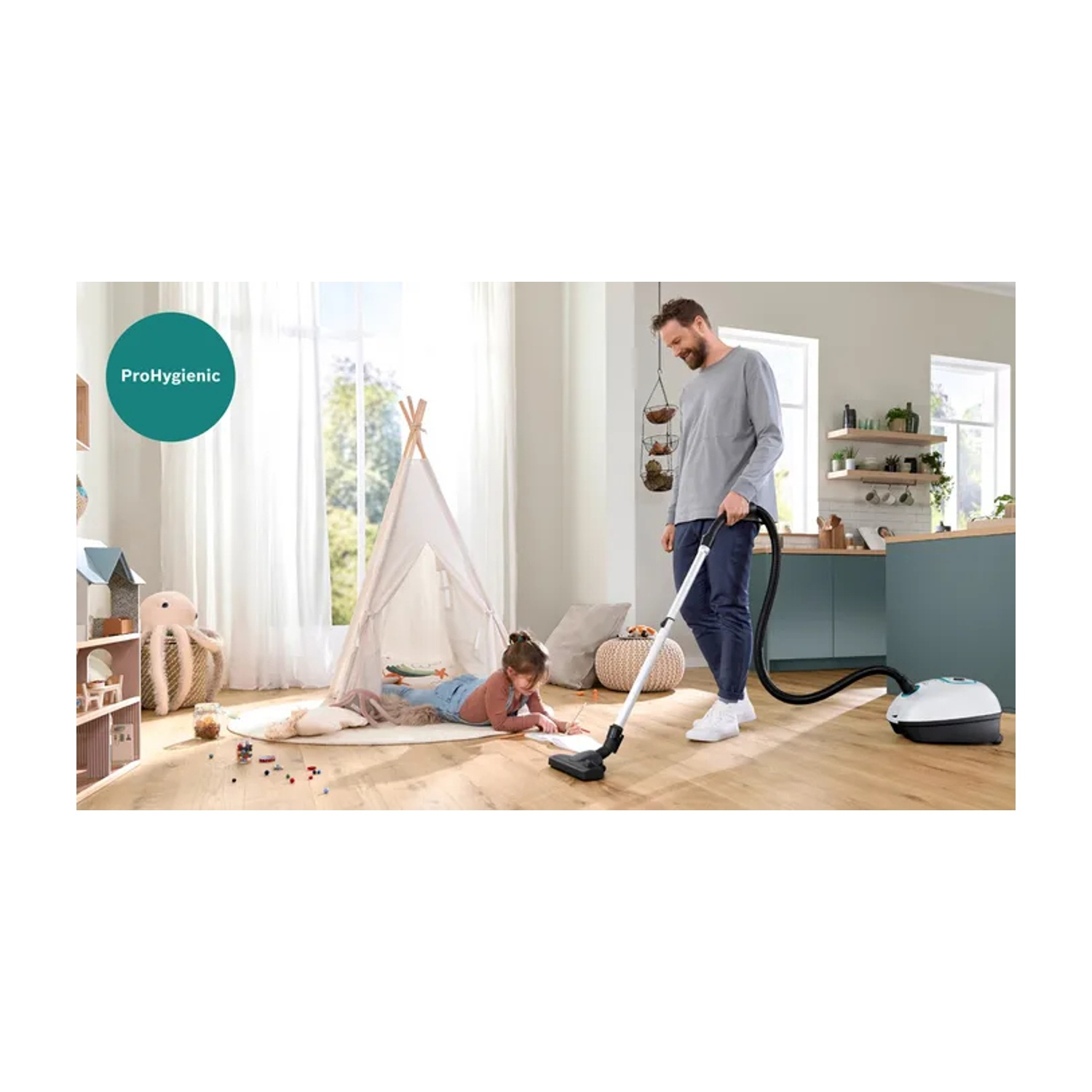 Un uomo aspira una stanza con un bambino che disegna sul pavimento. Un teepee e giocattoli sullo sfondo.