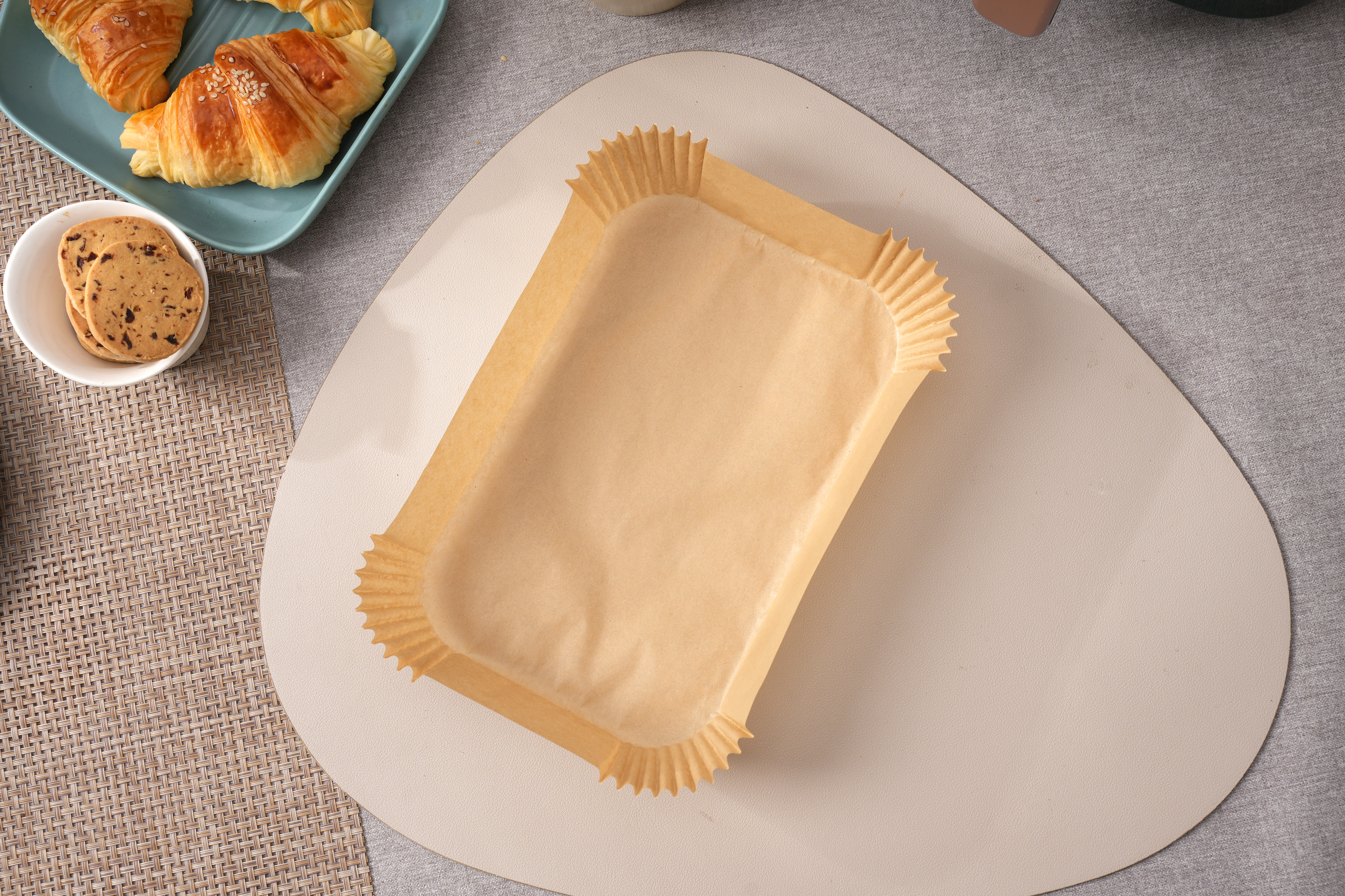 Brązowa papierowa forma do pieczenia na jasnoszarej powierzchni. Rogaliki i ciasteczka w tle.