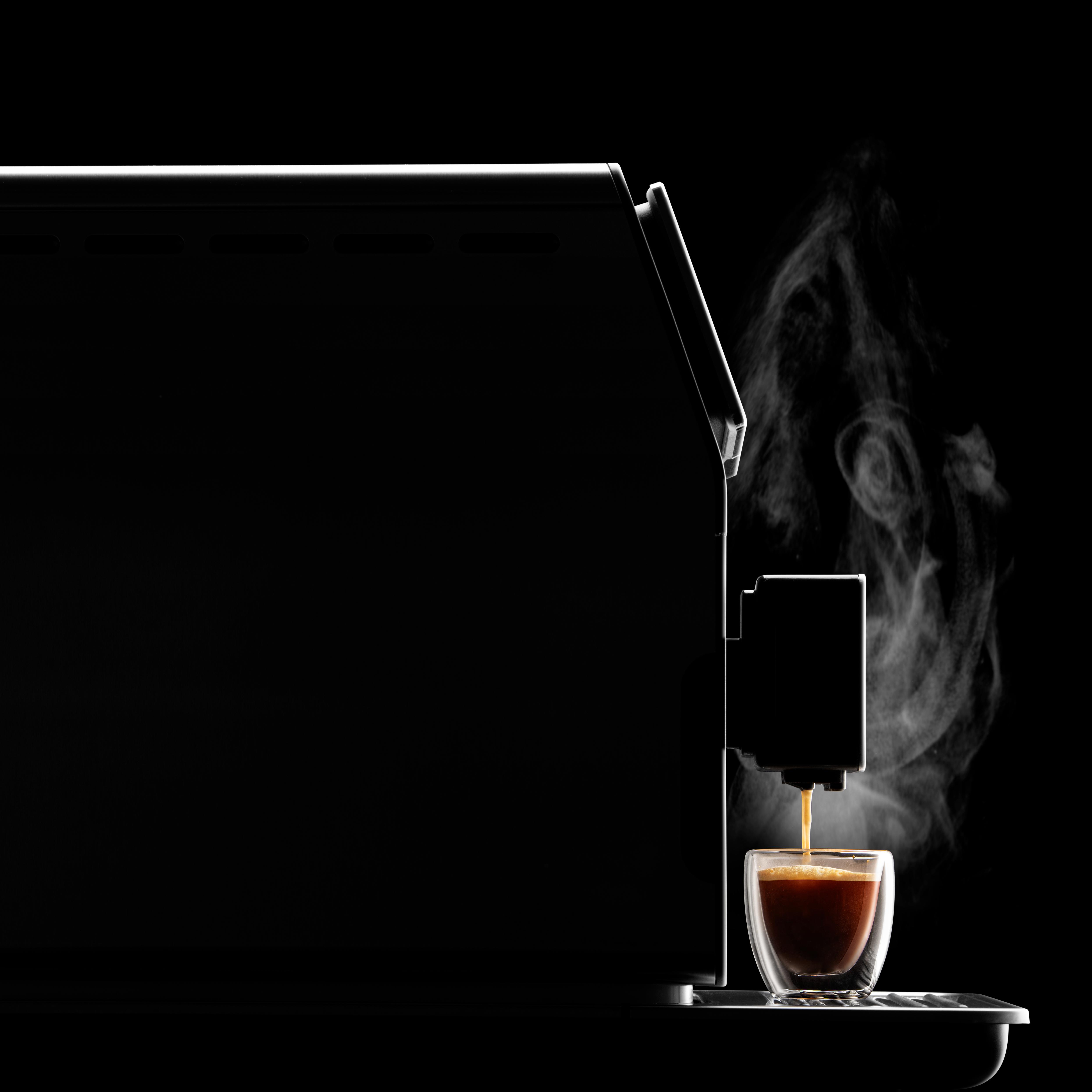 Kaffeemaschine gießt Kaffee in ein Glas. Dampf und dunkler Hintergrund.