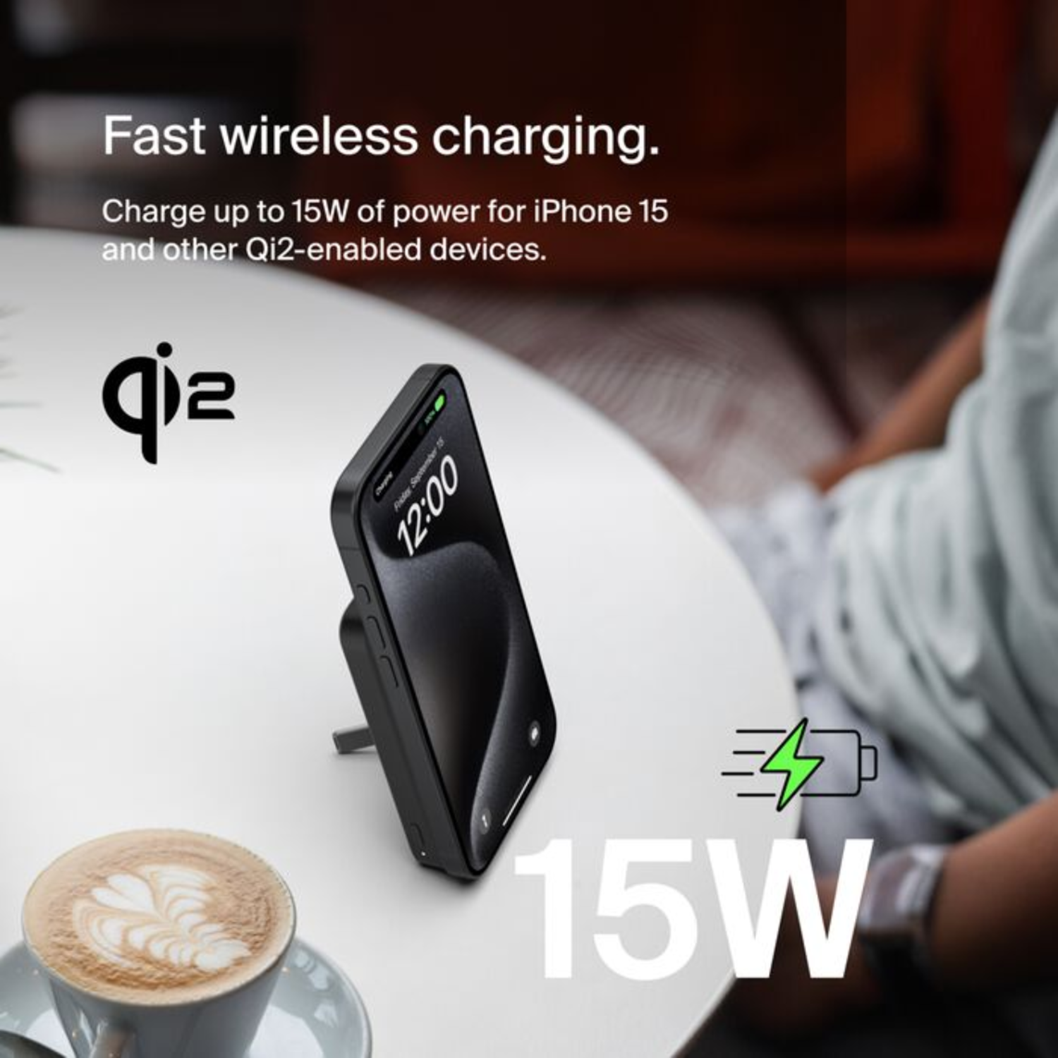 Téléphone noir sur chargeur, table blanche avec tasse à café. Symbole de charge de 15W et logo Qi2.