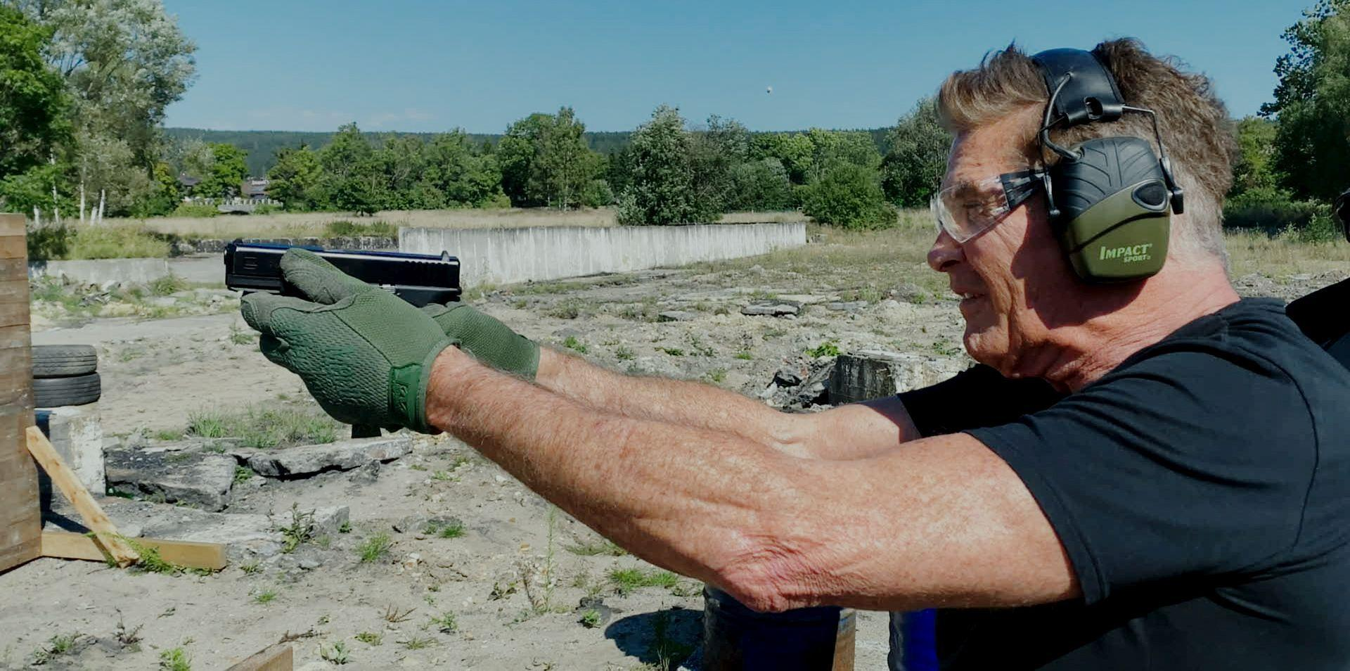 David Hasselhoff mit Gehörschutz und Brille schießt mit einer Waffe.