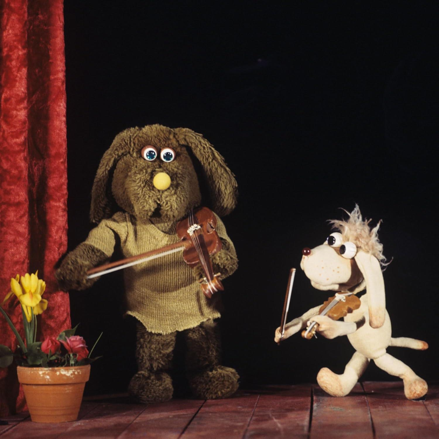 Zwei Puppen spielen auf einer Bühne Geige. Eine ist braun und pelzig, die andere beige und hundeartig.