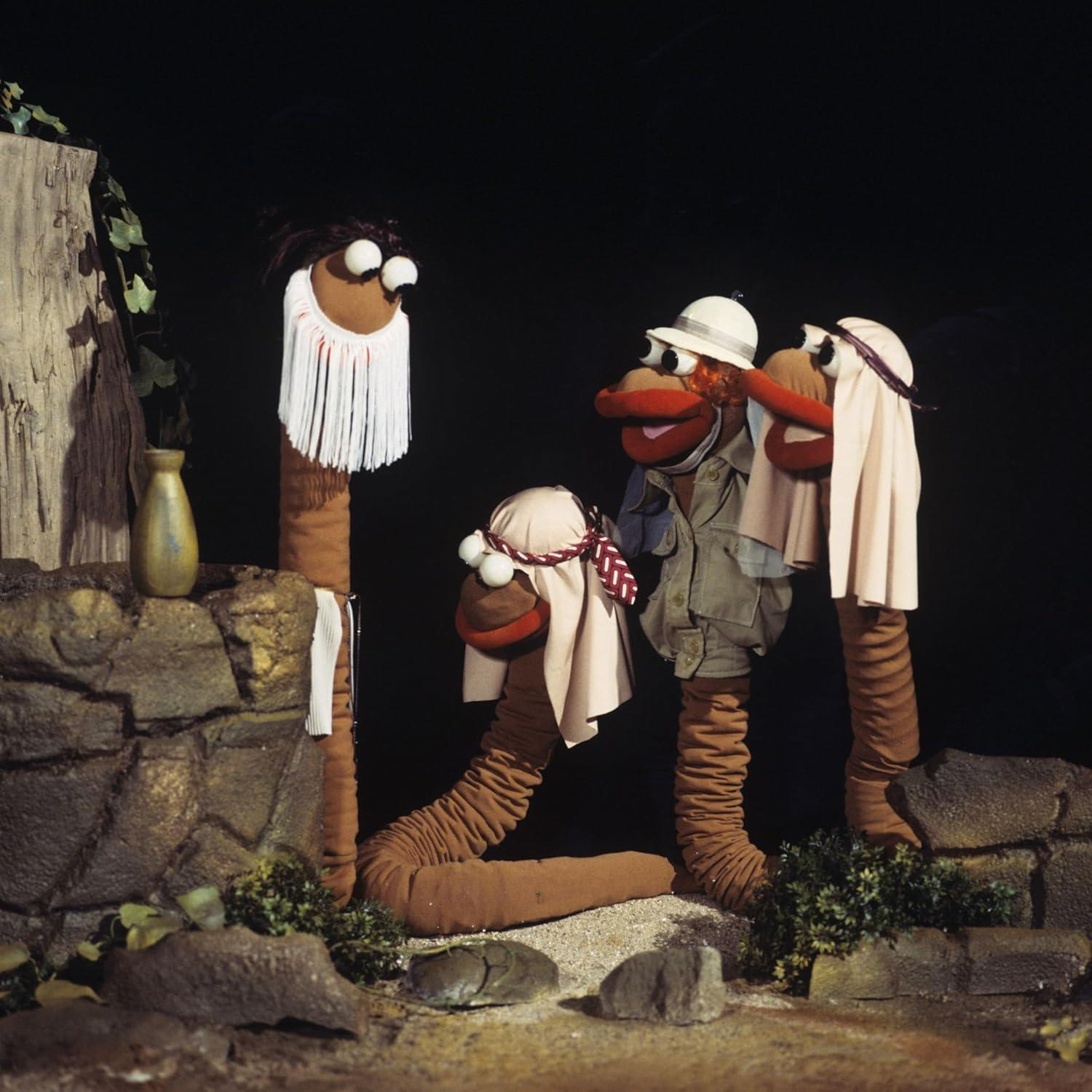 Drei wurmartige Puppen. Einer mit Bart, die anderen beiden mit Hüten. Sie stehen an einer Steinmauer.