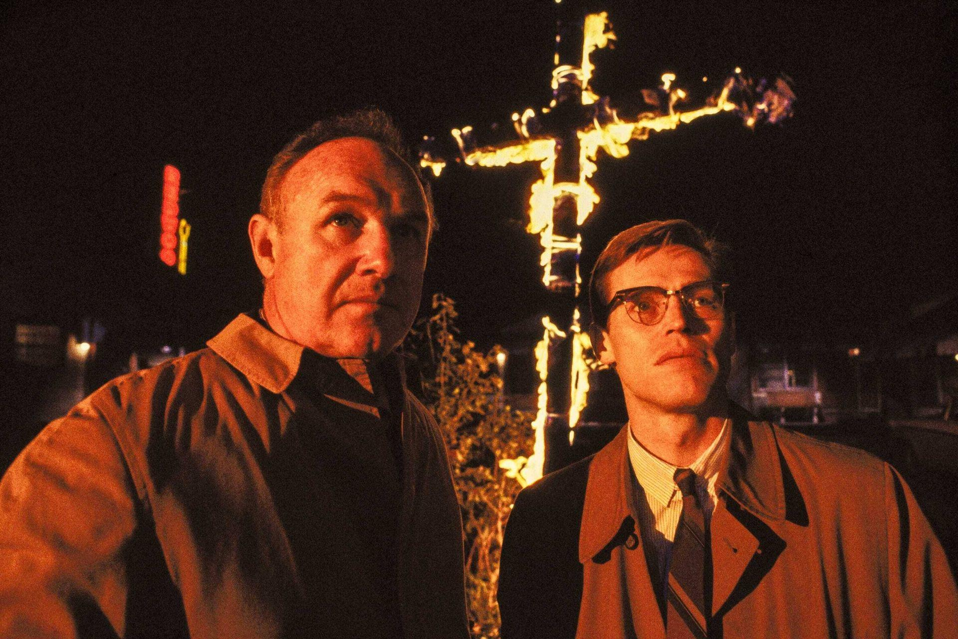 Zwei Männer stehen nachts vor einem großen, mit Lichtern beleuchteten Kreuz, im Hintergrund ein Neonschild.