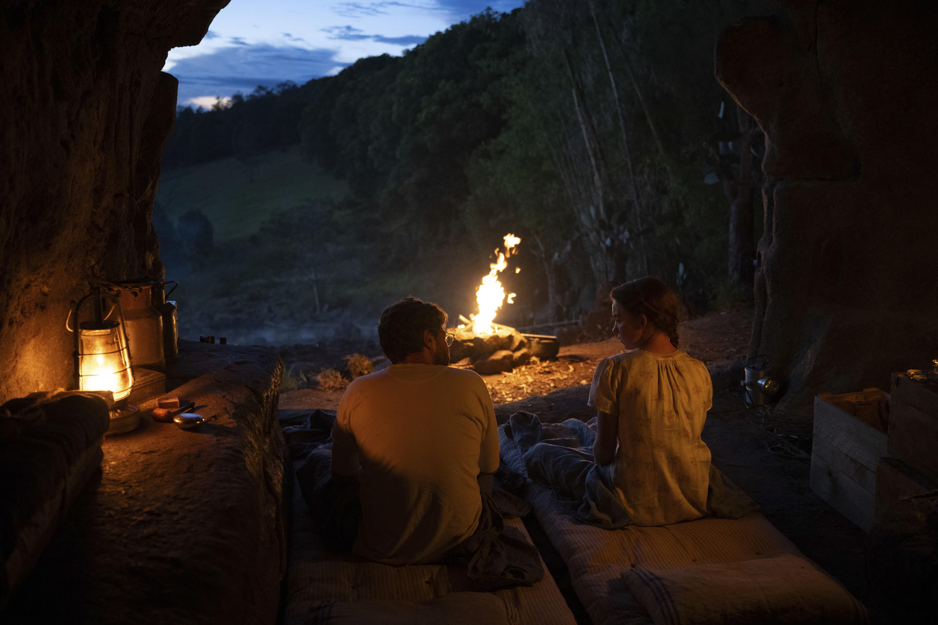 Zwei Personen sitzen bei einem Feuer in einem Höhleneingang bei Dämmerung. Laternen hängen, im Hintergrund ein Wald.