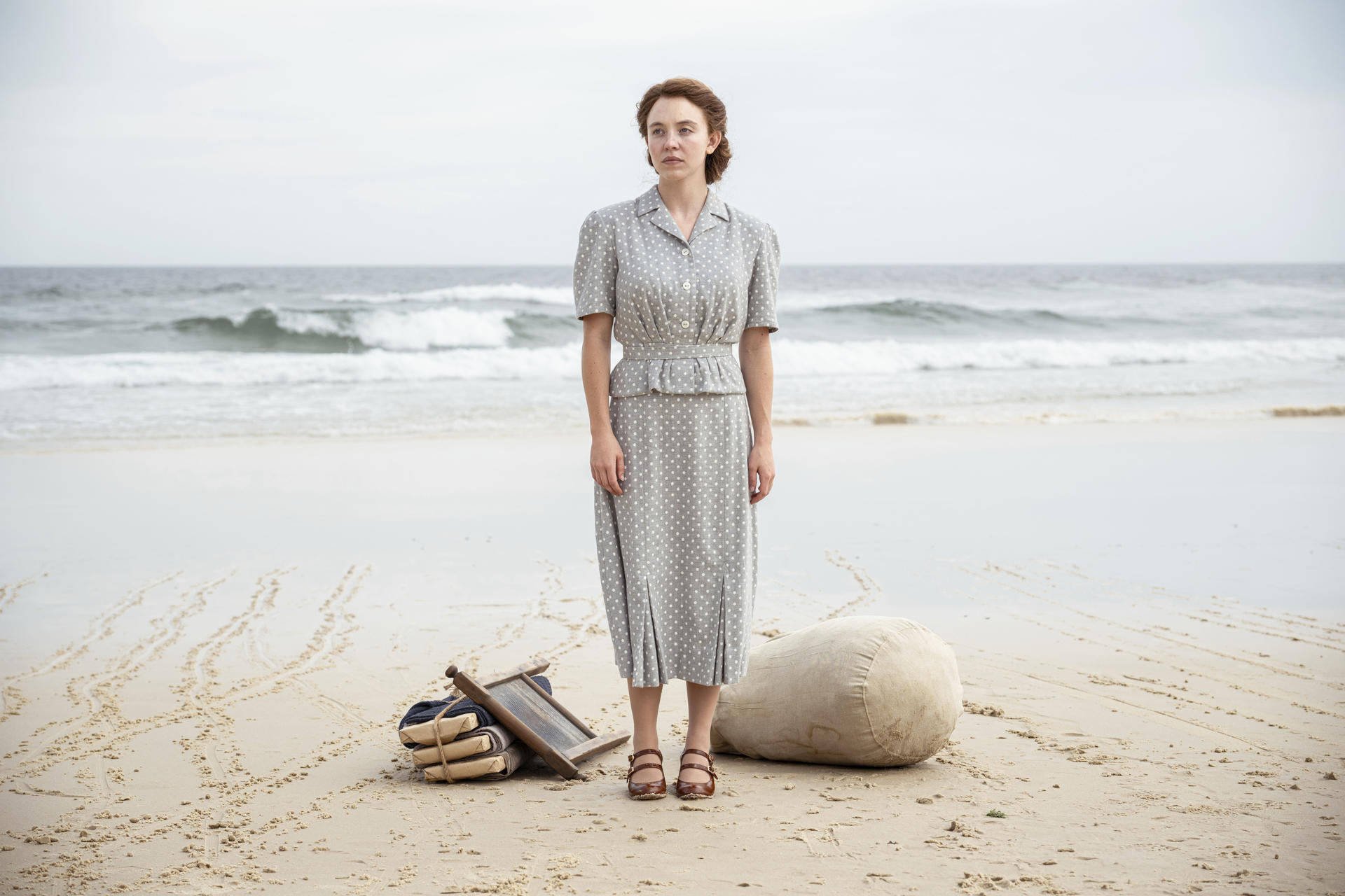 Frau am Strand, trägt ein Kleid und Sandalen, Blick zum Ozean. Strandutensilien in der Nähe.
