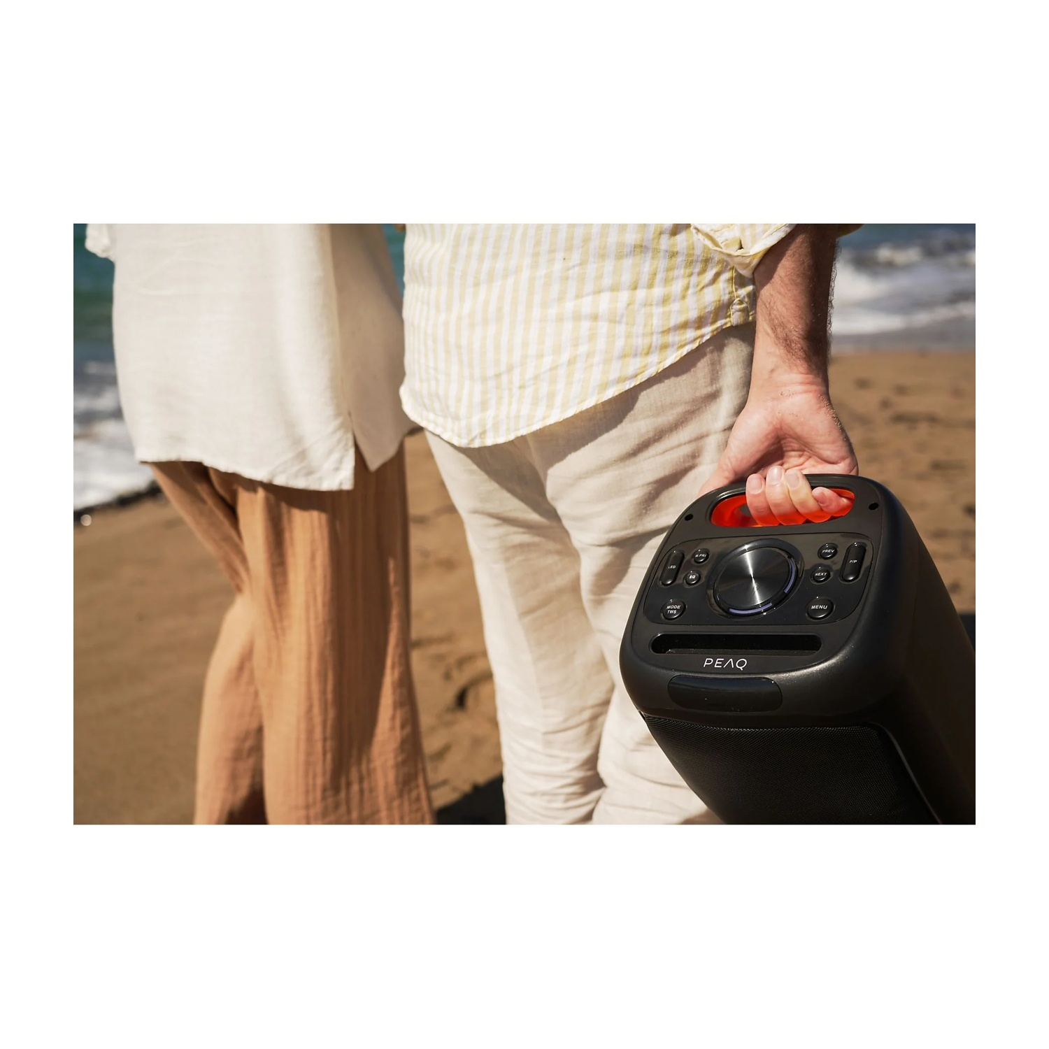 Uomo che tiene un altoparlante nero sulla spiaggia. Due persone sono in piedi vicino.