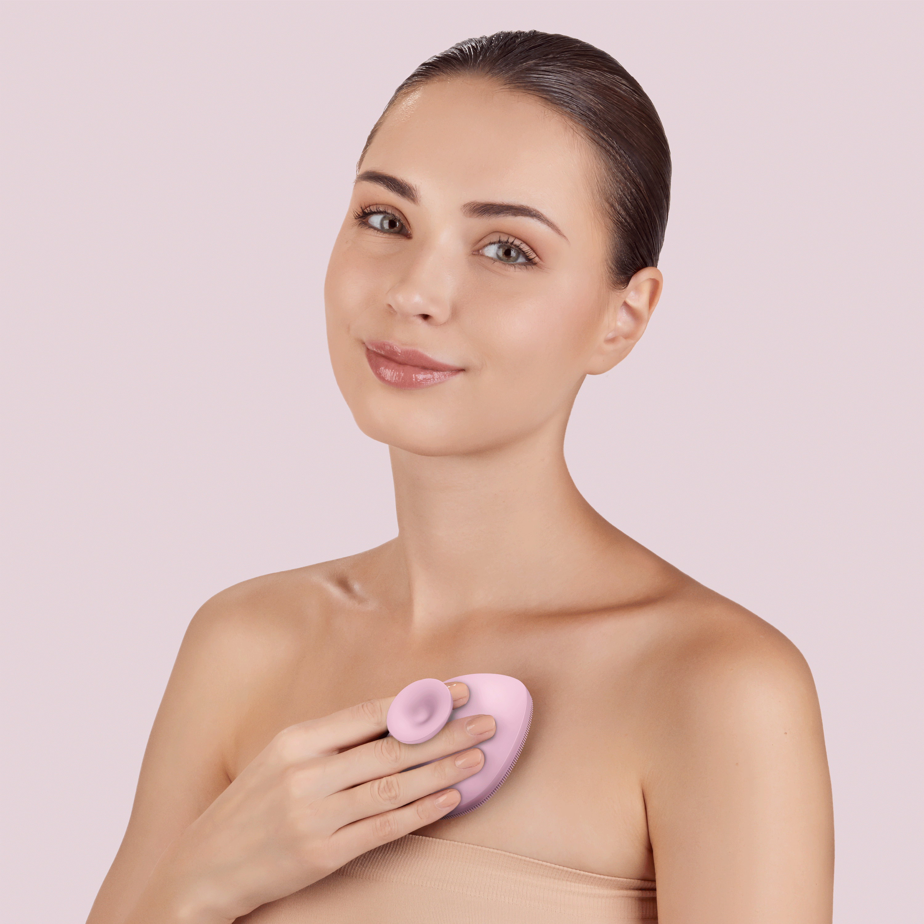 Bir kadın, göğsünün üzerinde pembe bir Geske SmartAppGuided vücut fırçası tutuyor. Pembe arka plan.