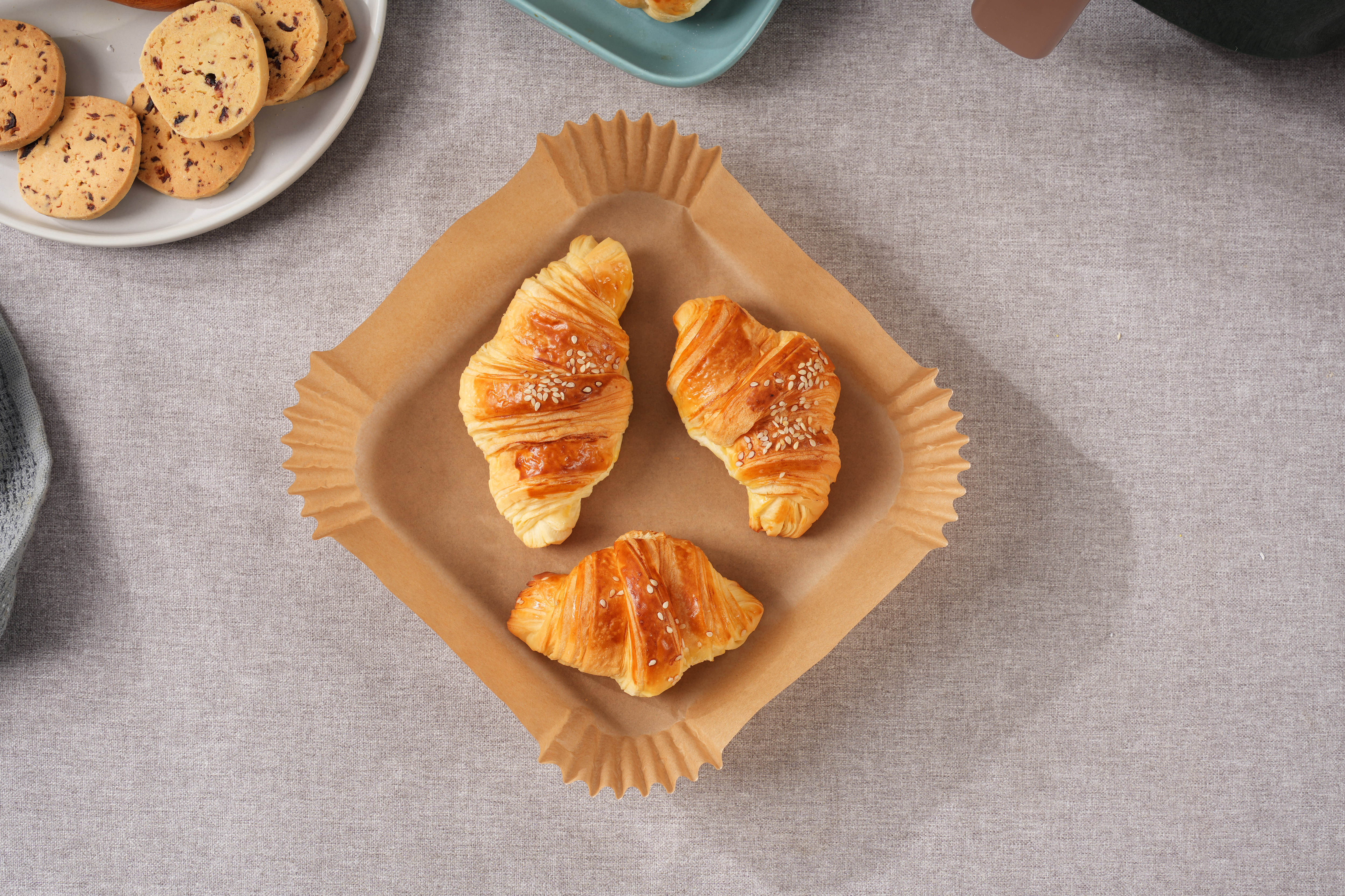 Drei Croissants in einer quadratischen, braunen Backform, neben einem Teller mit Keksen.
