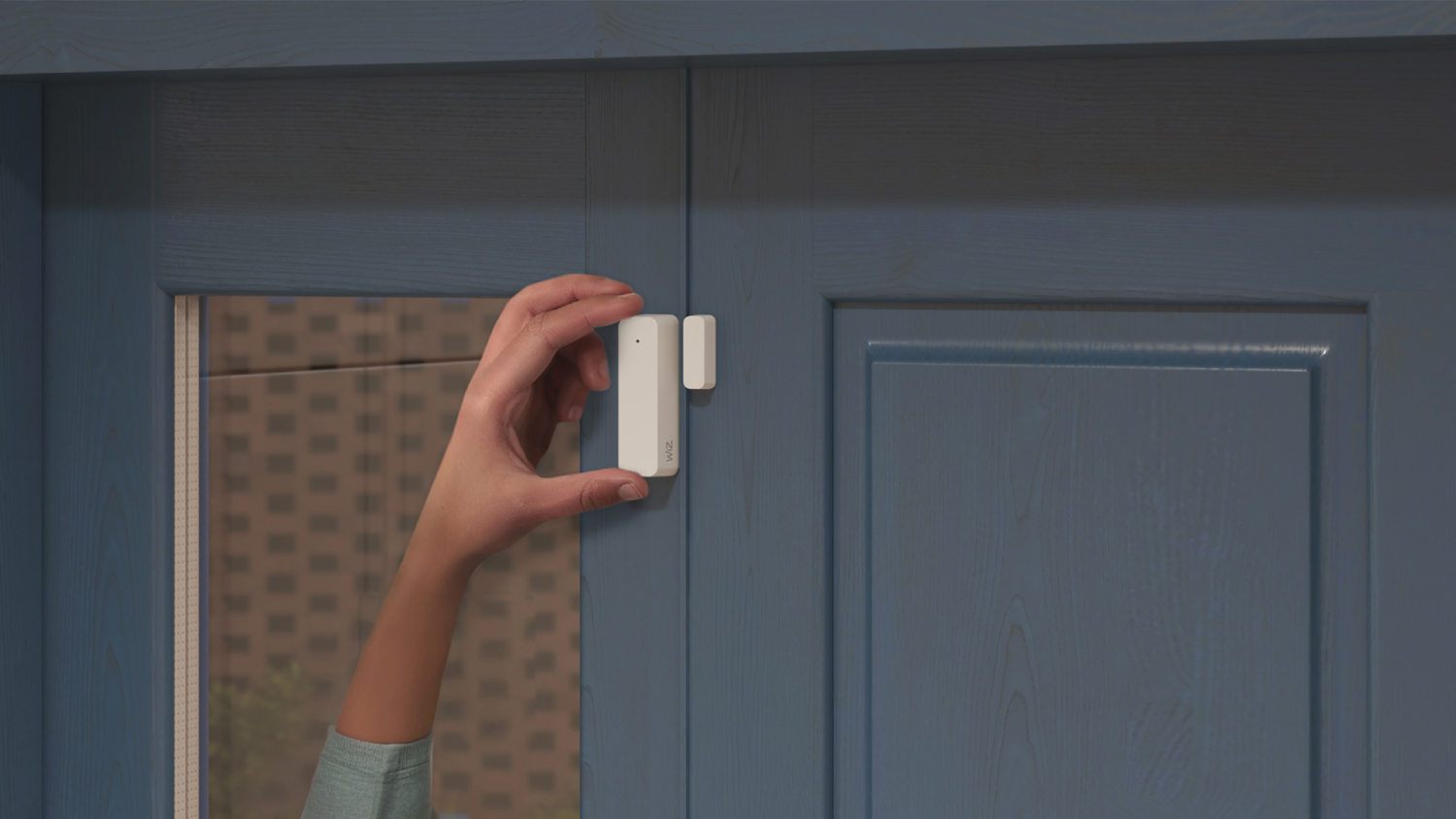 Una mano sostiene un sensor de contacto Wiz blanco en una puerta azul.
