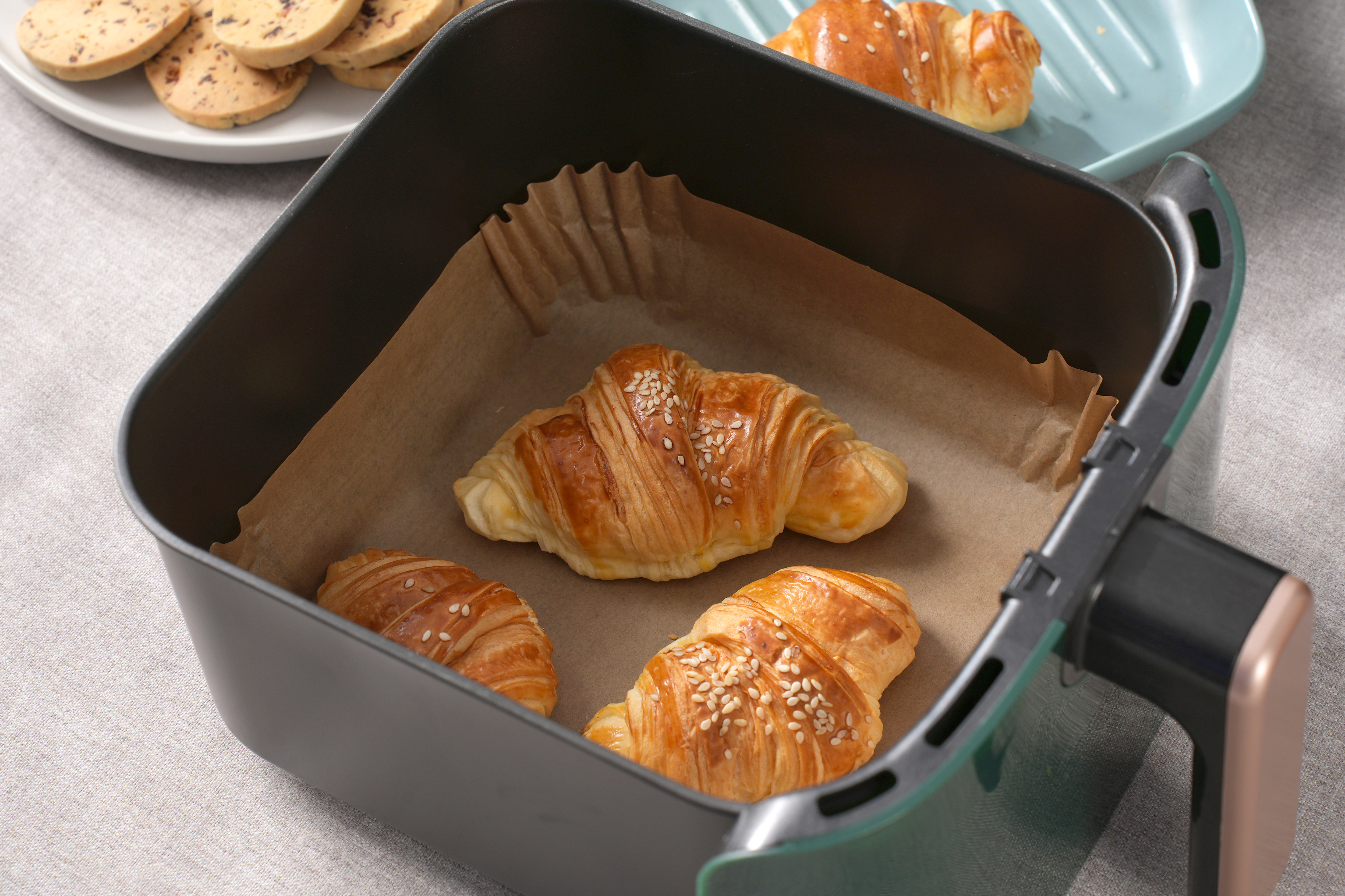 Croissants in einem schwarzen Airfryer-Korb, neben einem Teller mit Keksen. Braunes Pergamentpapier.