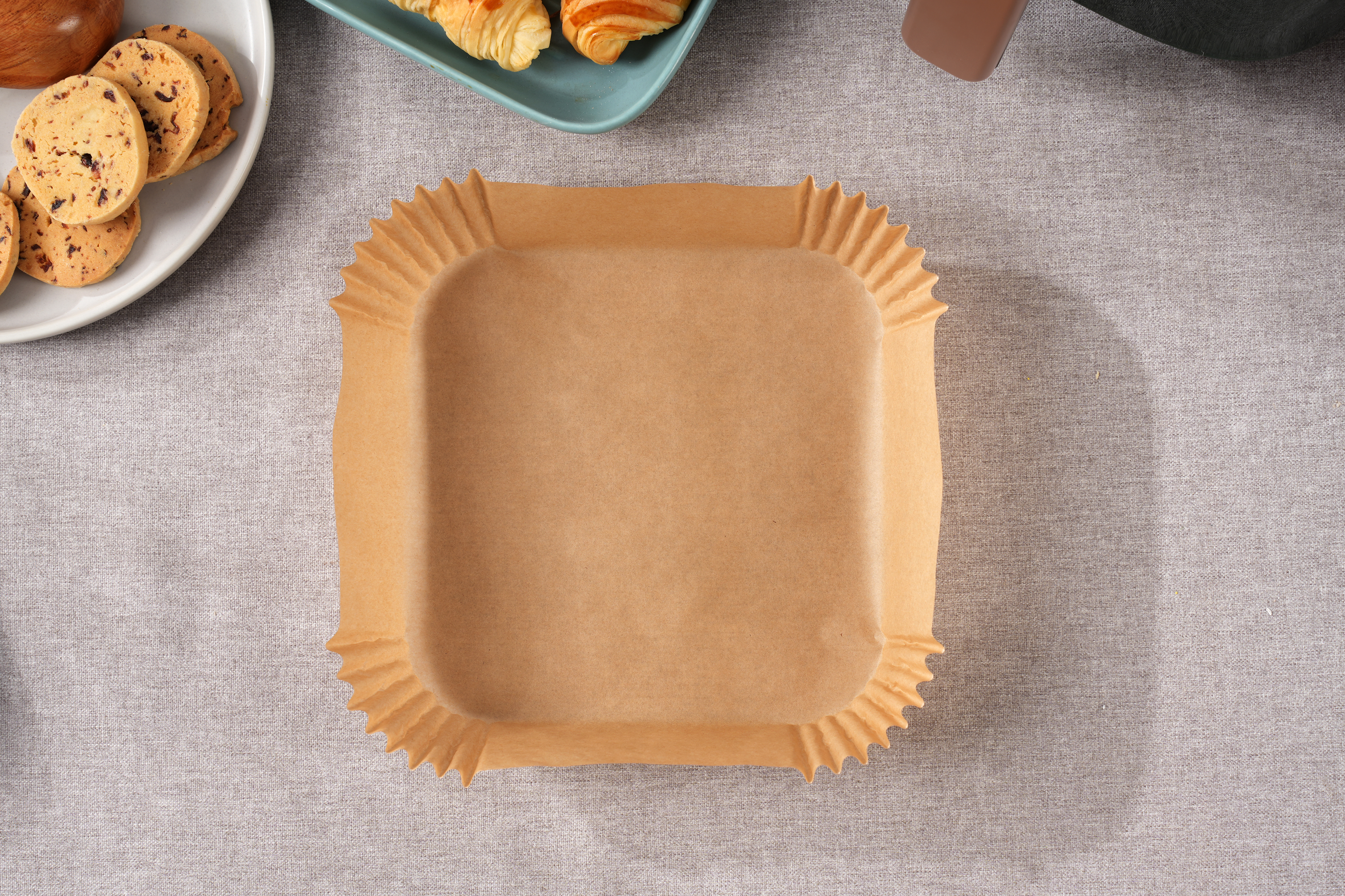 Eine braune Papierbackform ist zentriert. Kekse und Croissants befinden sich im Hintergrund auf einer grauen Tischdecke.